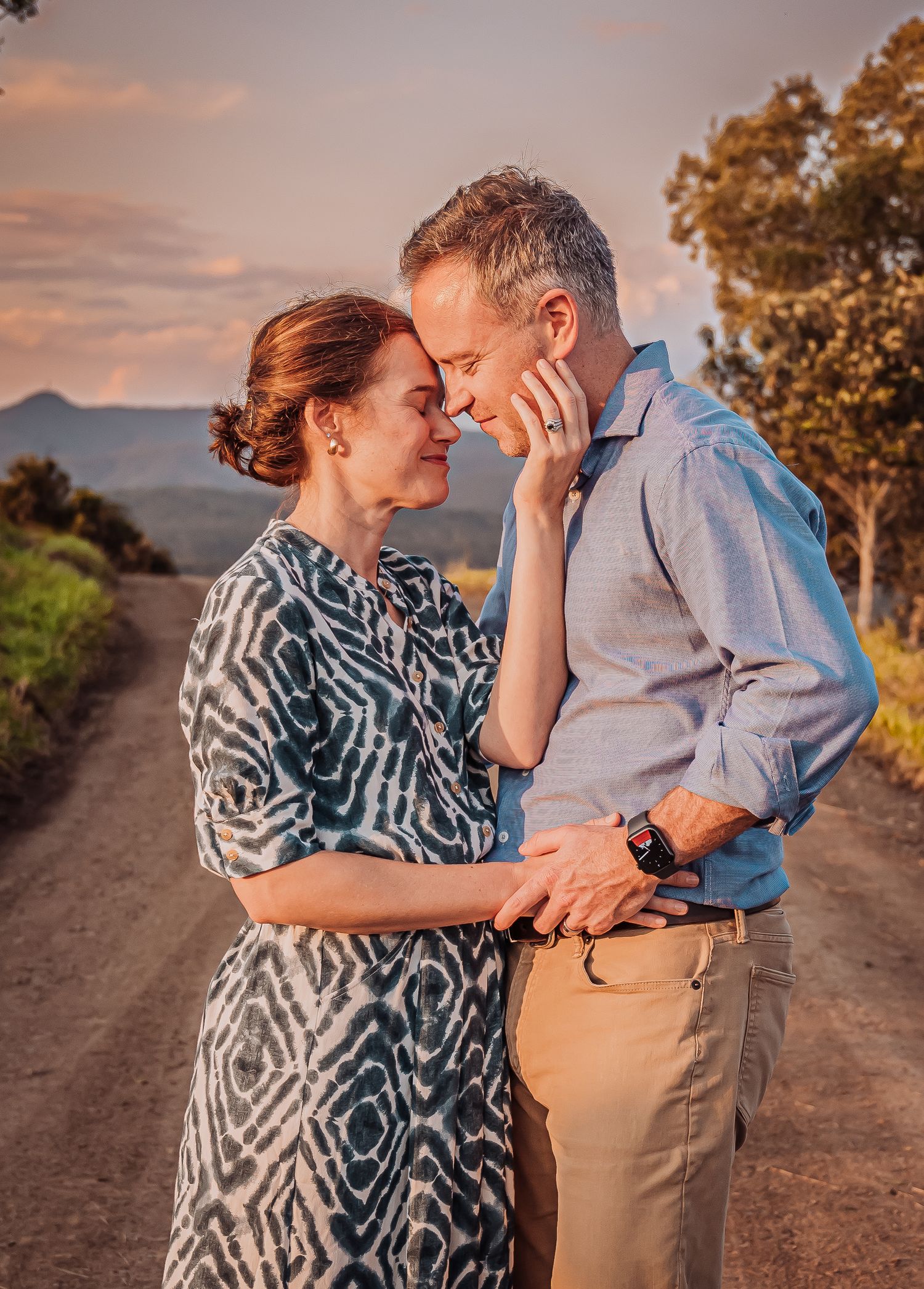 A romantic couple embraces on a rural road during golden hour with mountains visible in the distance.