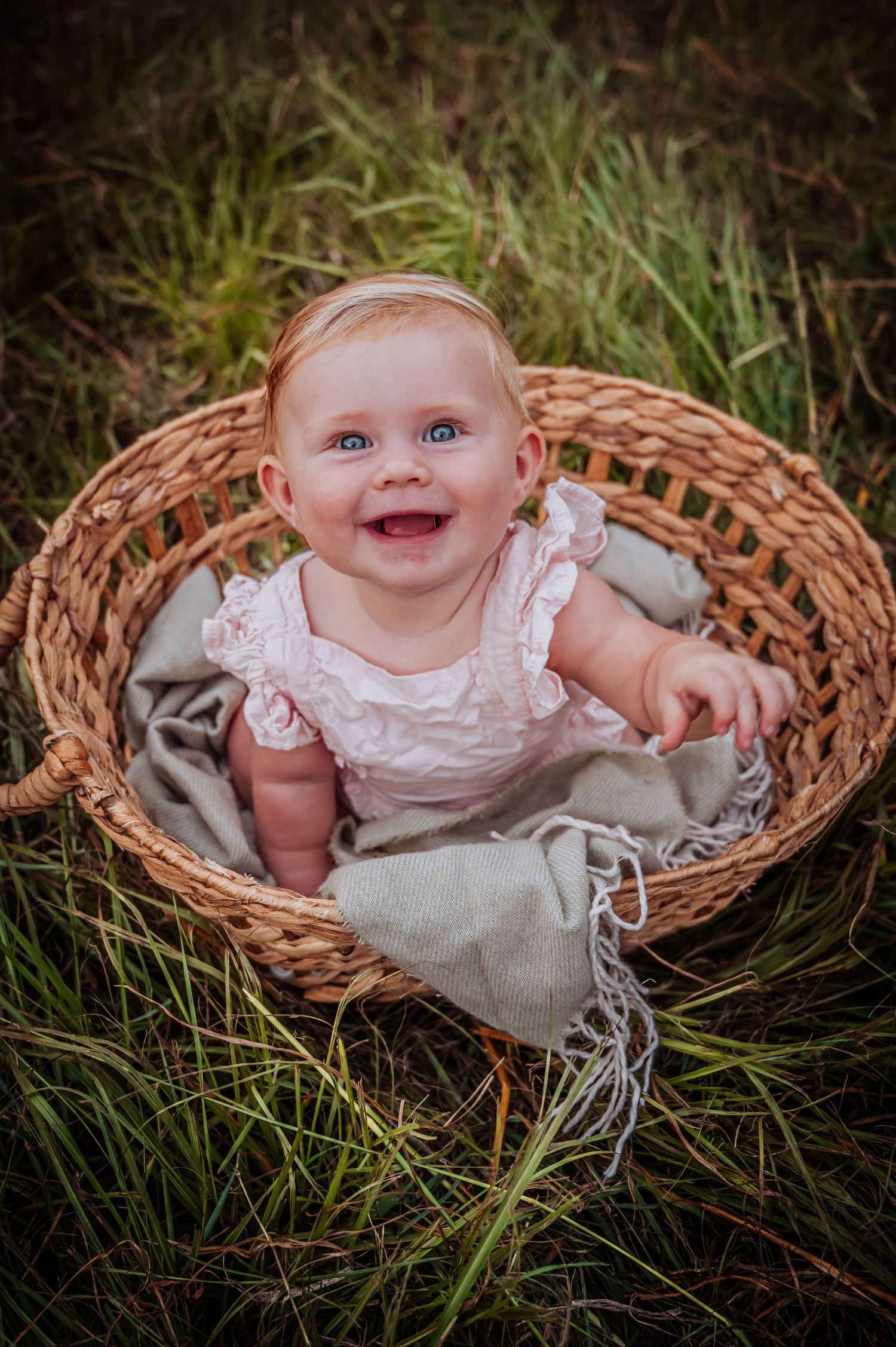 Happy baby sitting in woven basket wearing white lace dress outdoors in grass.