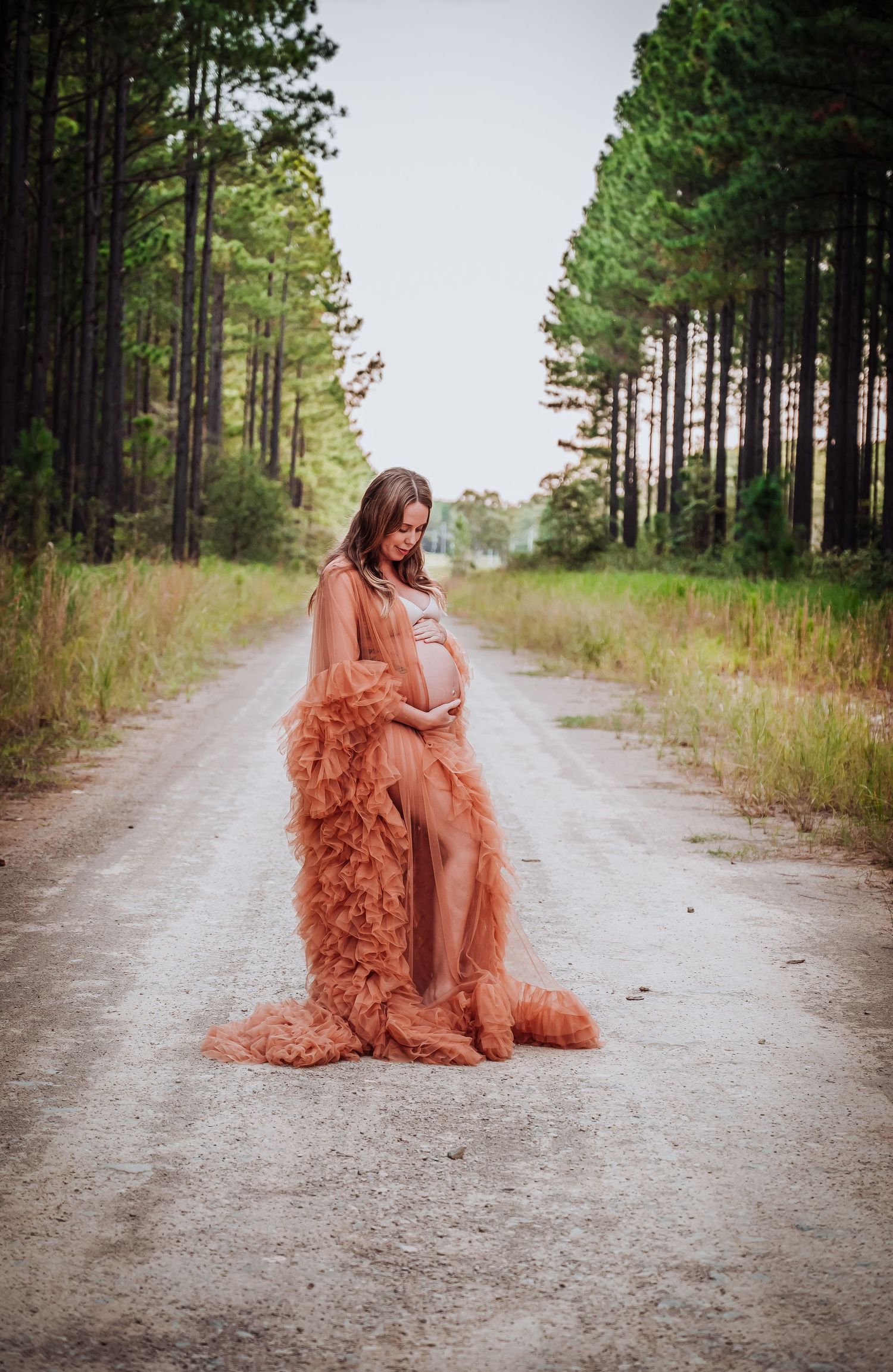 A maternity portrait on a tree-lined dirt road featuring a flowing peach dress and natural lighting.