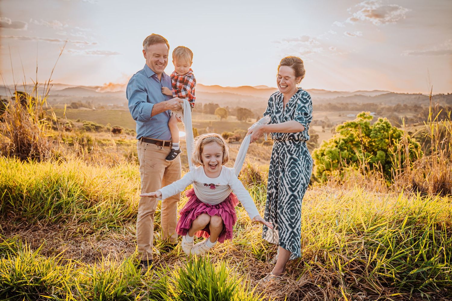 Family plays together in grassy field during golden hour with mountains in background.
