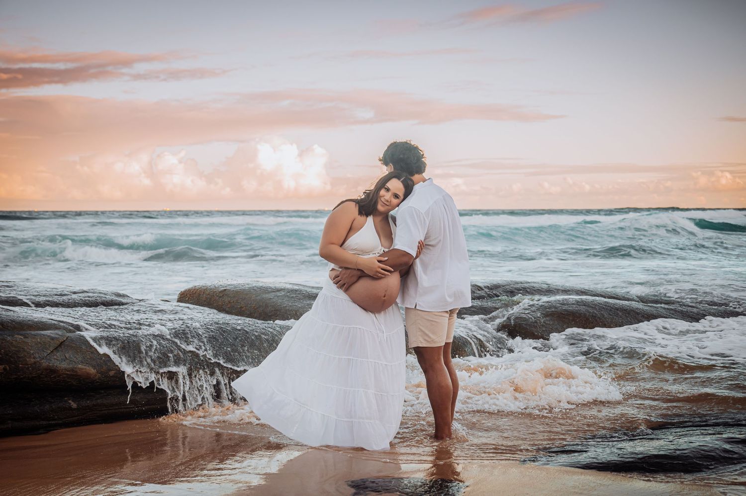 A romantic couple embraces on a rocky beach at sunset with crashing waves in the background.