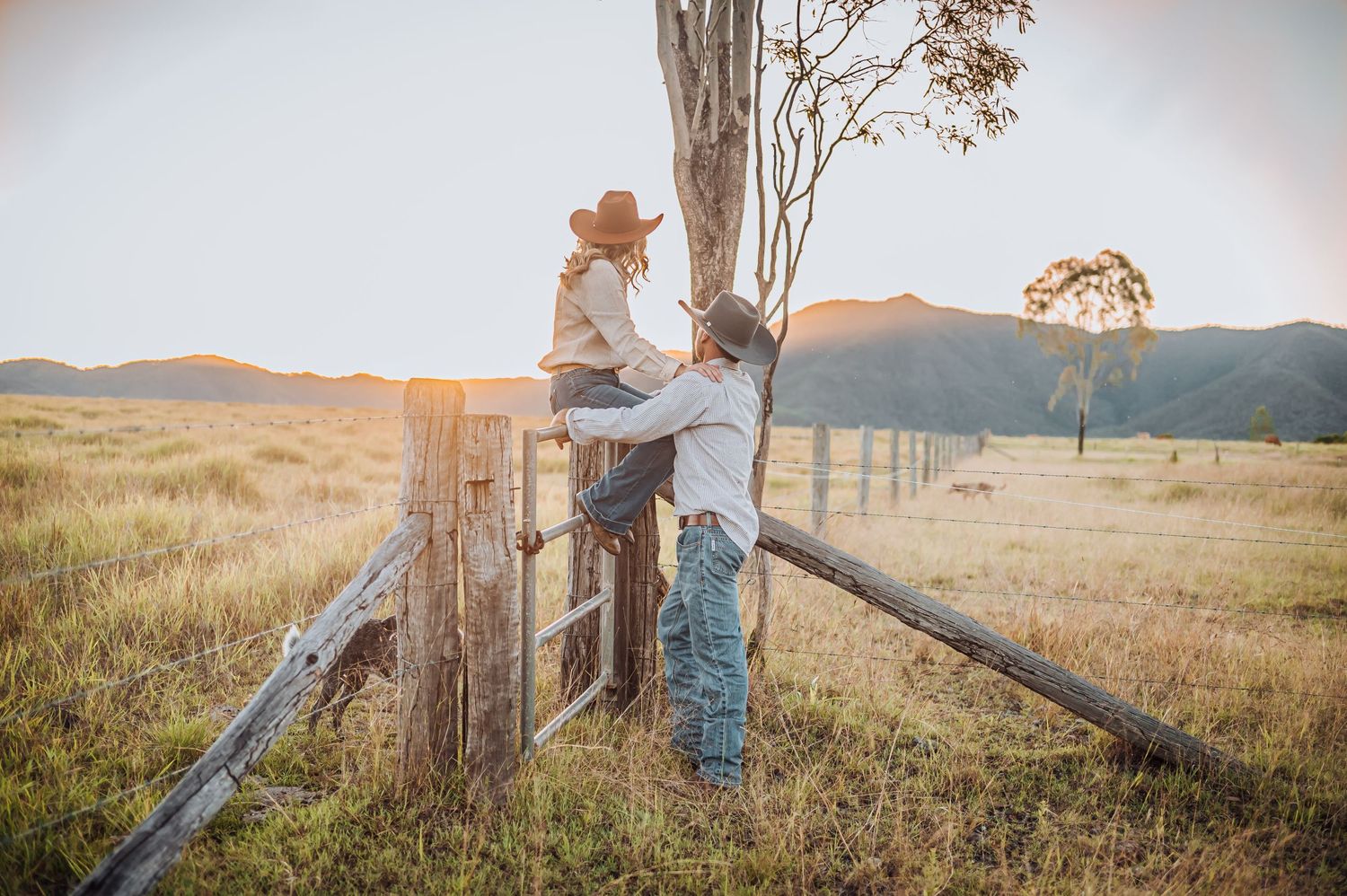 Two people embrace by an old wooden fence post at golden hour in a rural field.