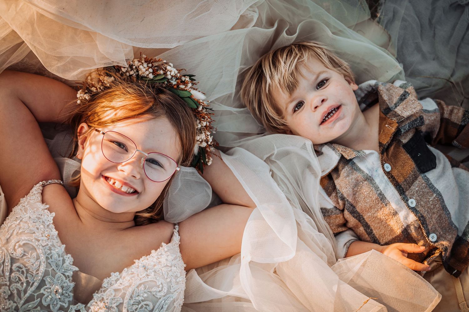 Two children lying together wearing floral crowns and formal attire in dreamy lighting.