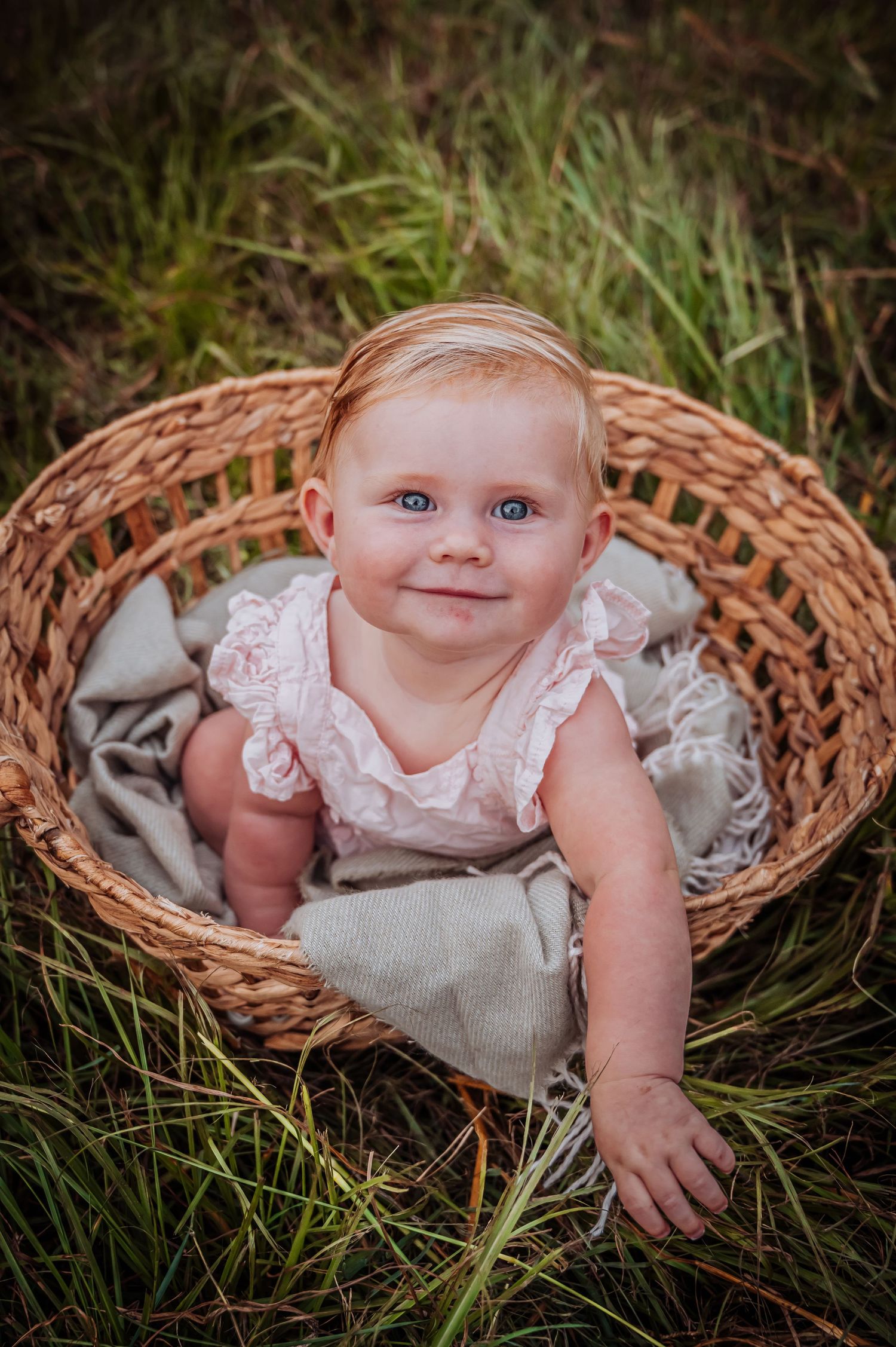Happy baby in white lace dress posing in rustic wicker basket outdoors.
