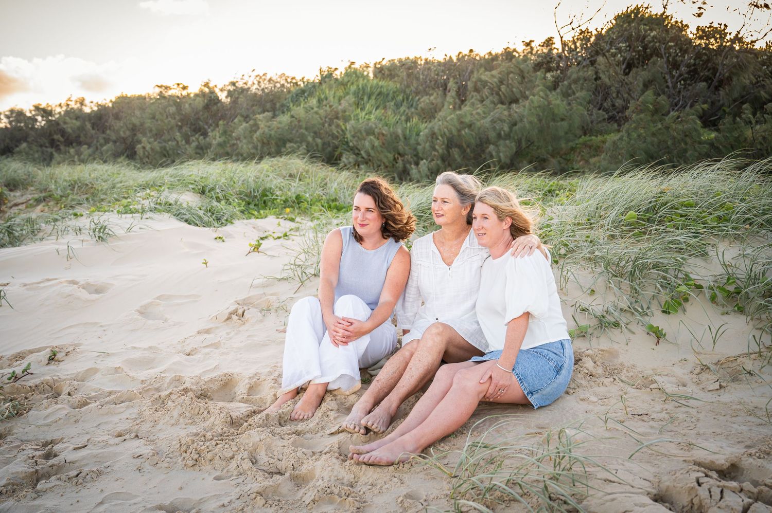 Three people sitting casually together on sand dunes at sunset.