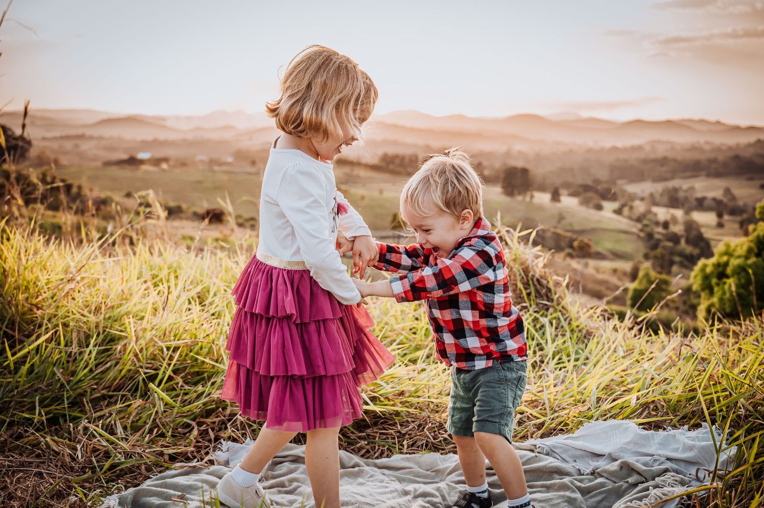 Two young children in summer outfits playing together on a hilltop with scenic rolling hills in background.