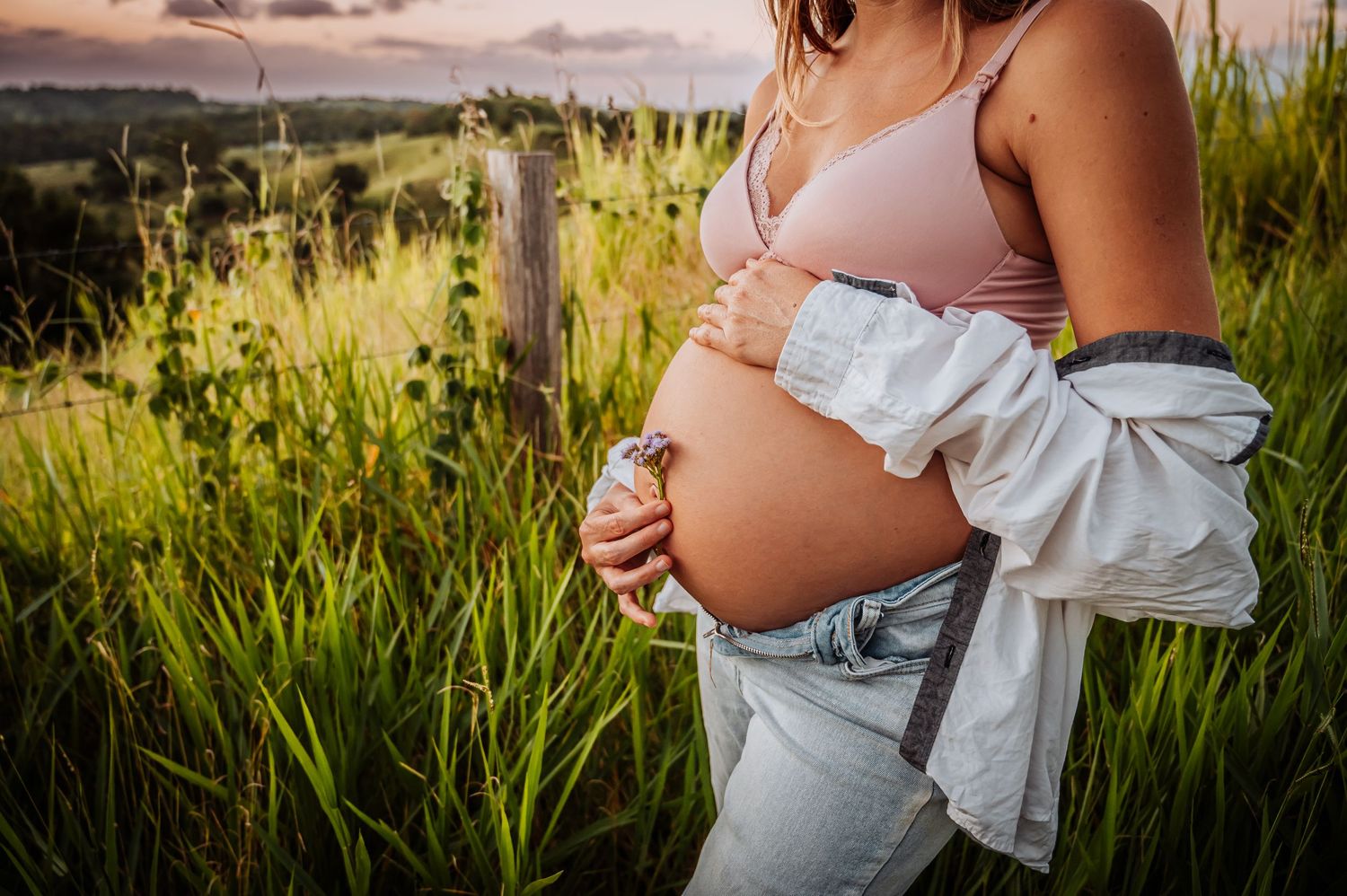 Intimate maternity photo showing pregnant belly against backdrop of sunlit field at dusk.
