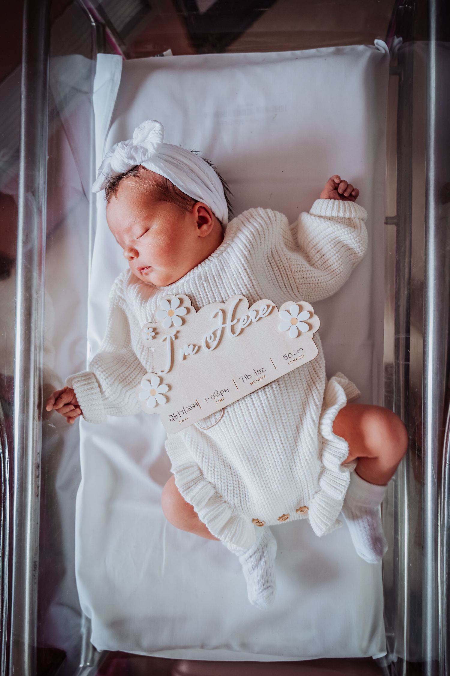 Newborn baby sleeping in clear hospital bassinet wearing white ruffled outfit.