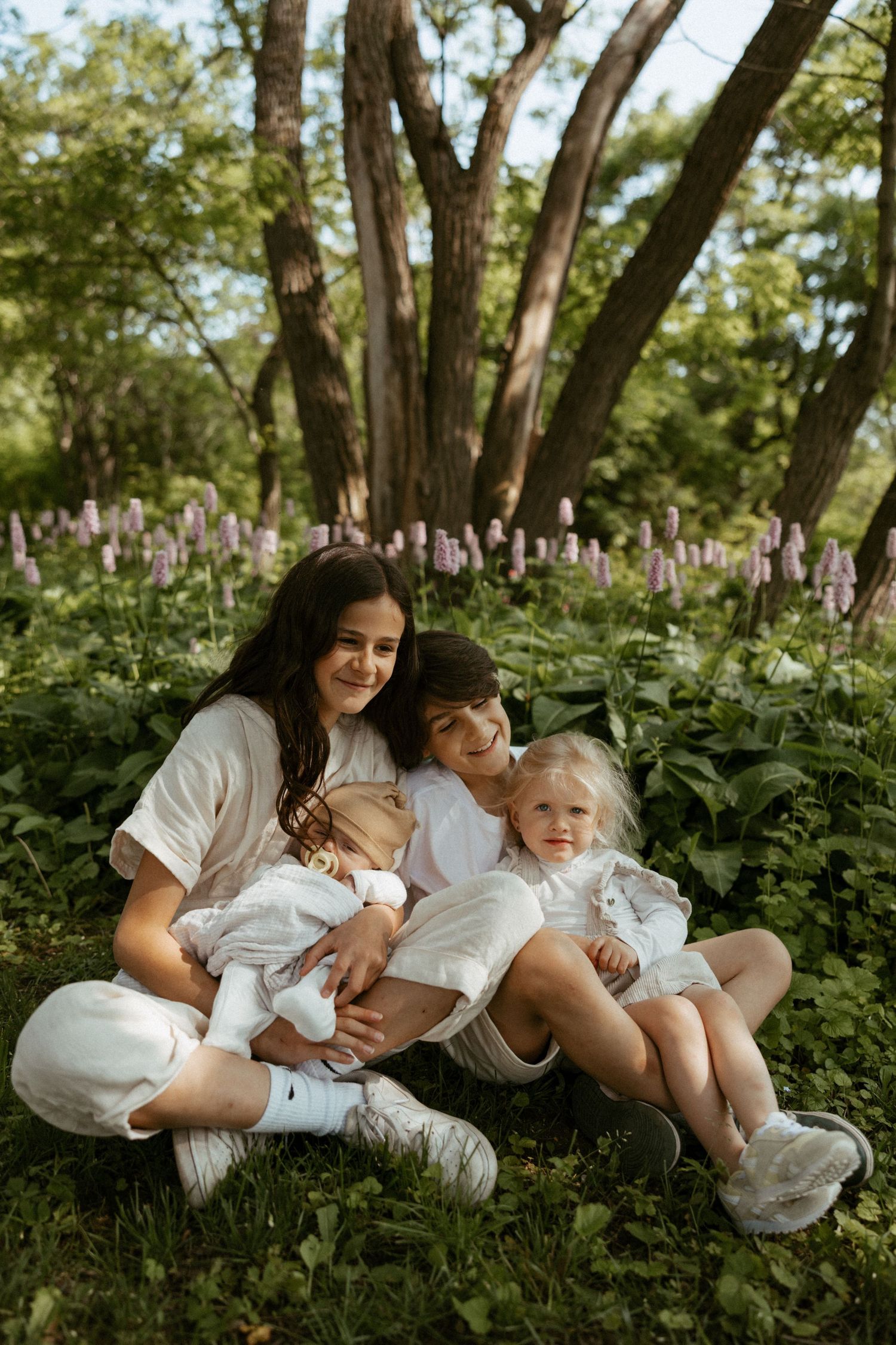 A mother sits on grass with three children in white outfits, surrounded by green trees and purple wildflowers.