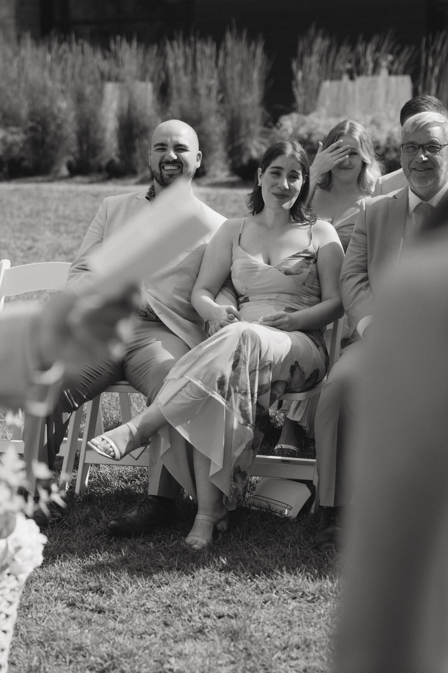 Black and white photo of smiling wedding guests seated outdoors on white chairs during a sunny garden ceremony.