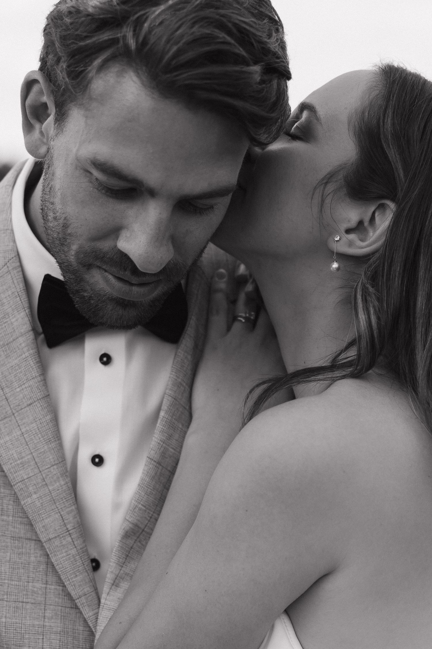 Black and white photo of a couple in wedding attire sharing an intimate moment, groom in bow tie suit.