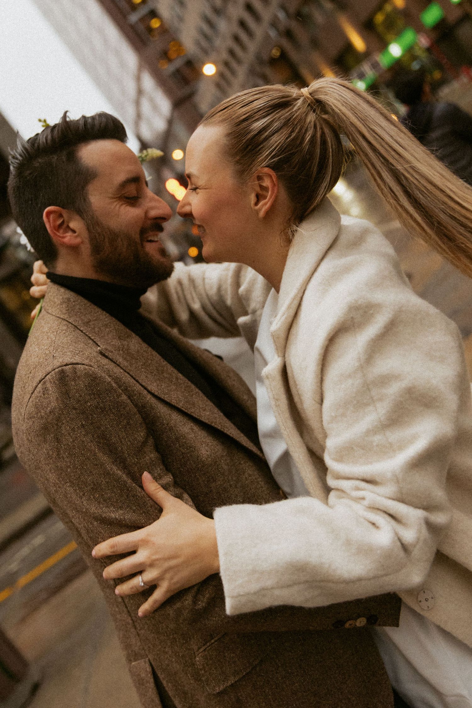 A couple embracing and smiling at each other outdoors, dressed in elegant coats with city lights in the background.