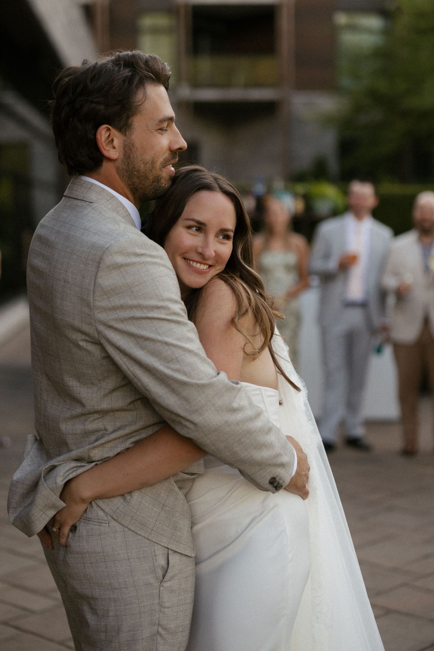 A smiling bride in a white dress embraces a groom in a gray suit outdoors at a wedding reception.