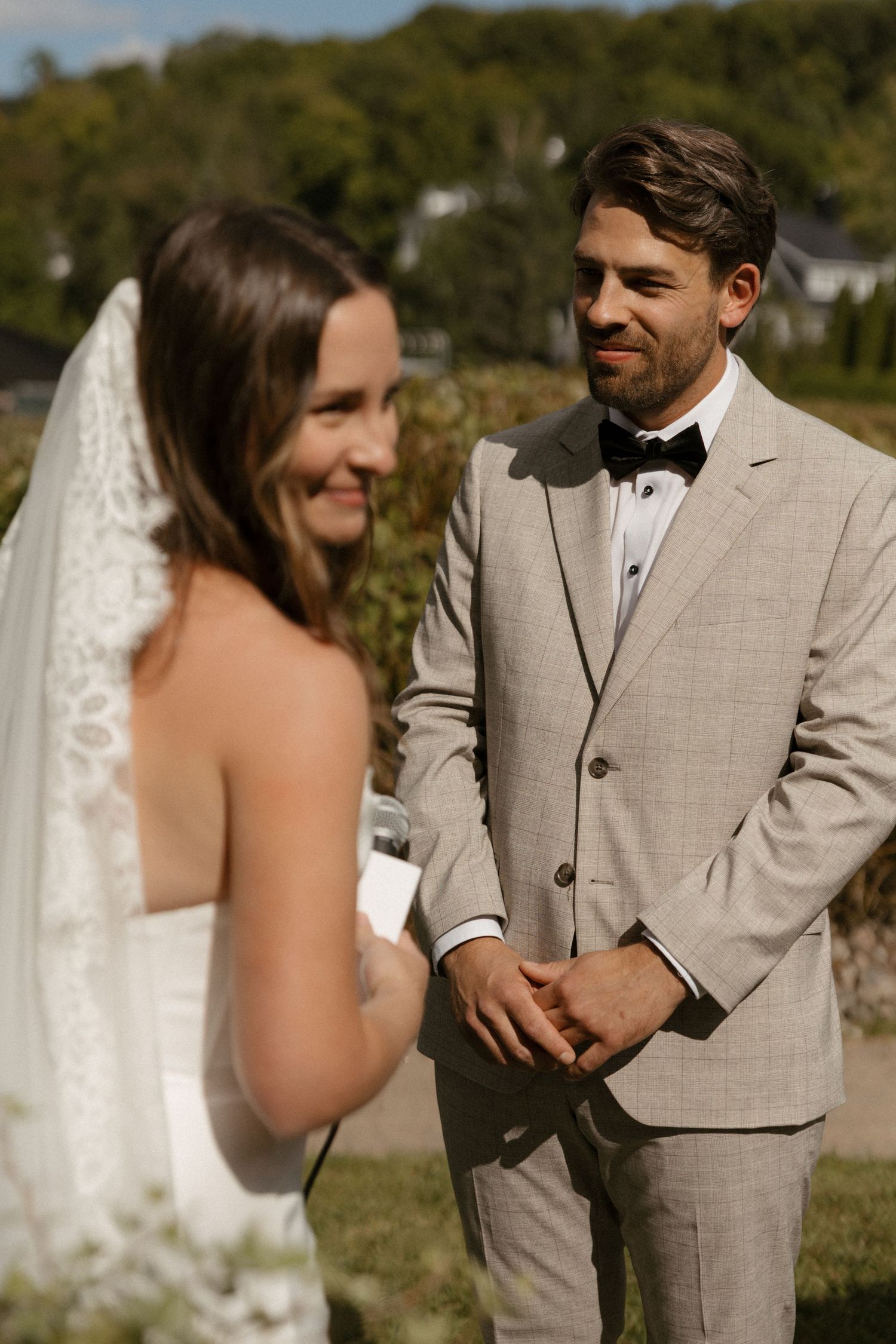 Groom in gray suit smiling at bride in white dress and veil during outdoor wedding ceremony.