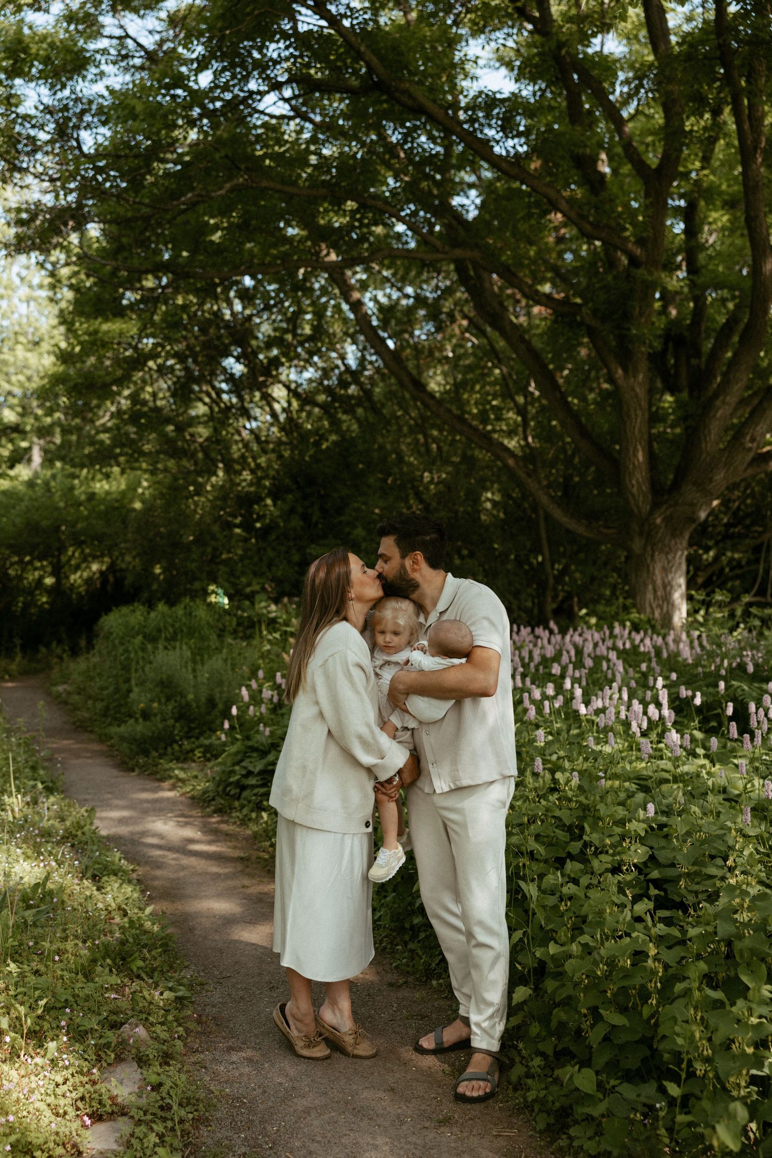 A young family in matching cream outfits shares a tender moment on a garden path surrounded by lush greenery.