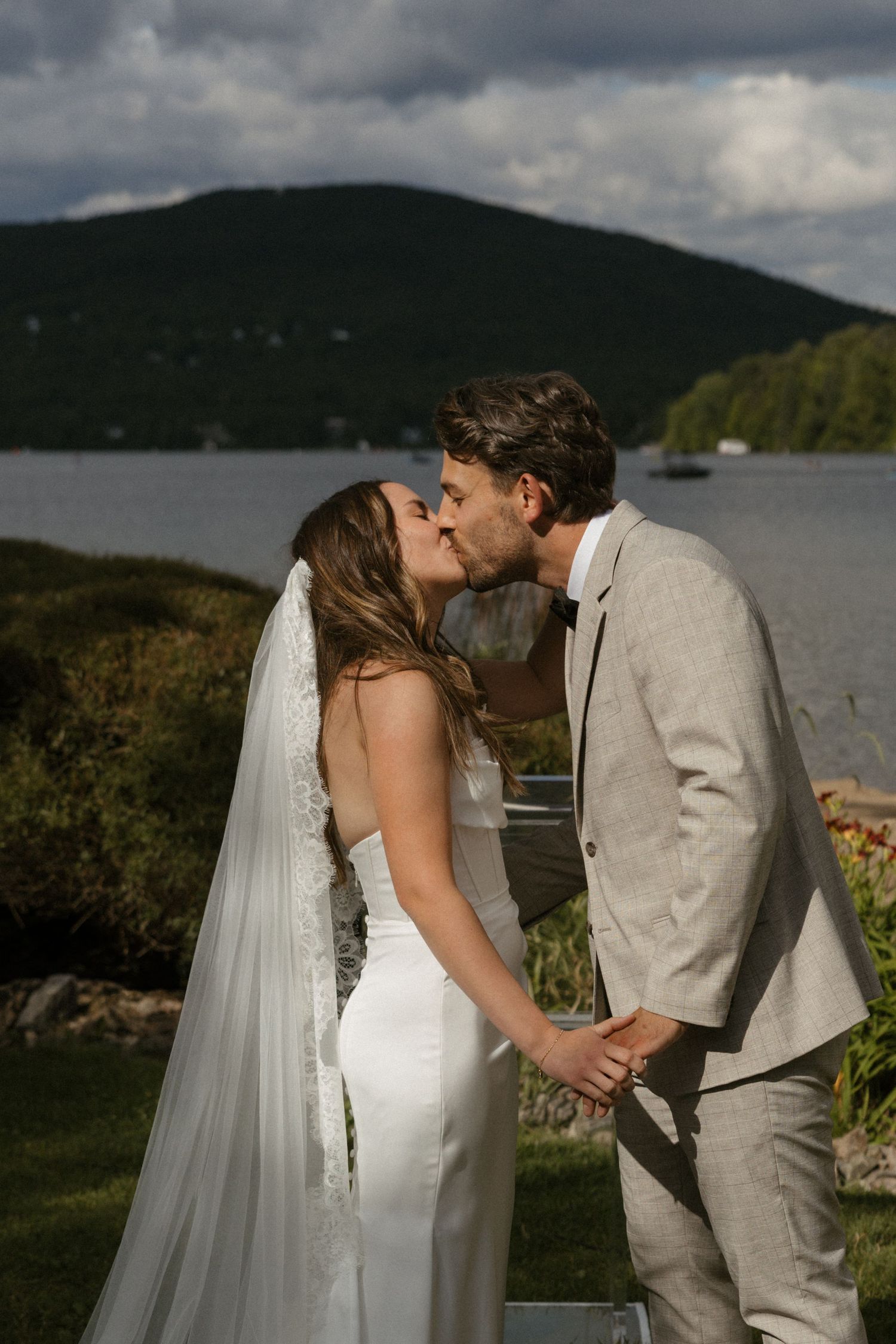 Bride and groom share a kiss at outdoor wedding ceremony with scenic lake and green hills in background.