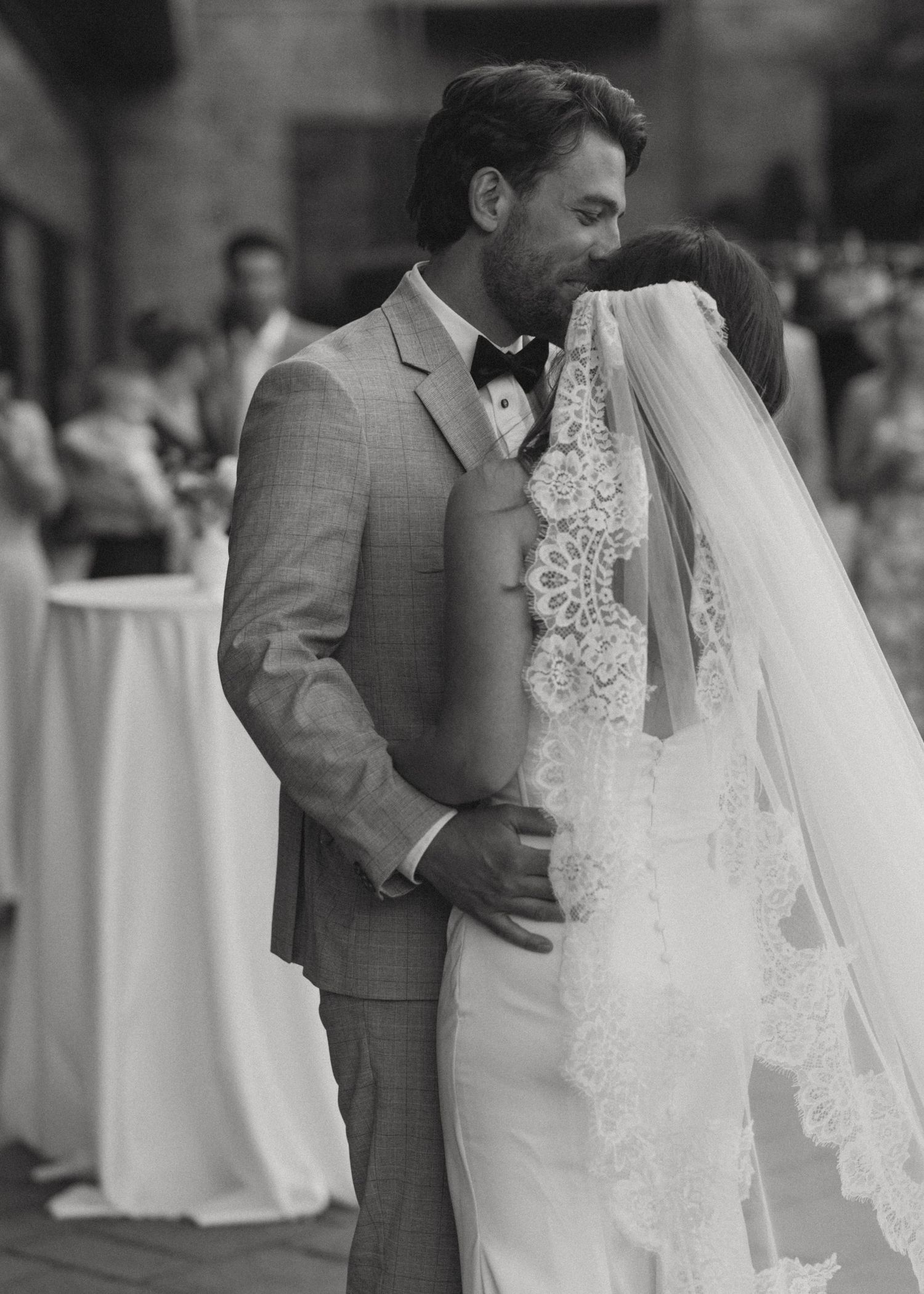 Black and white photo of a groom in a gray suit kissing his bride in a lace veil at their wedding.