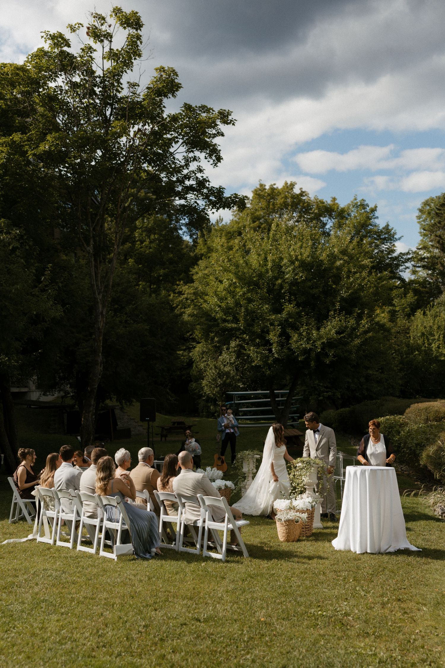 Outdoor wedding ceremony on green lawn with guests seated in white chairs facing bride and groom beneath lush trees.