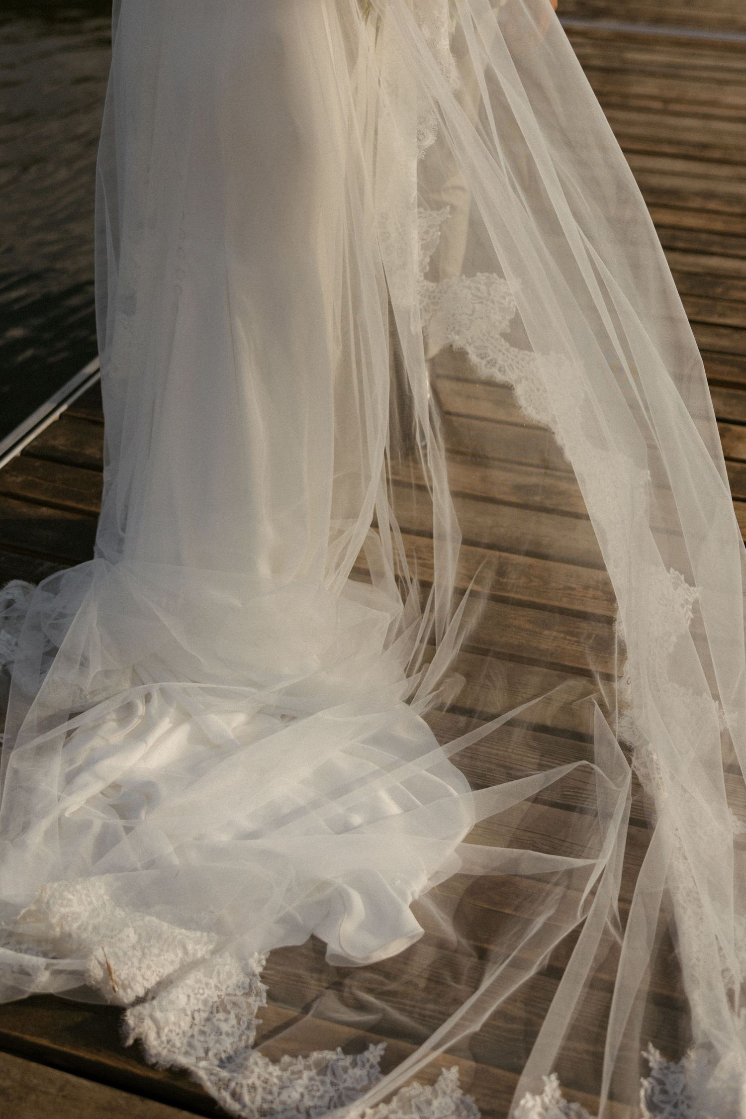 Elegant white wedding dress with lace-trimmed veil train spread across a wooden dock near water.