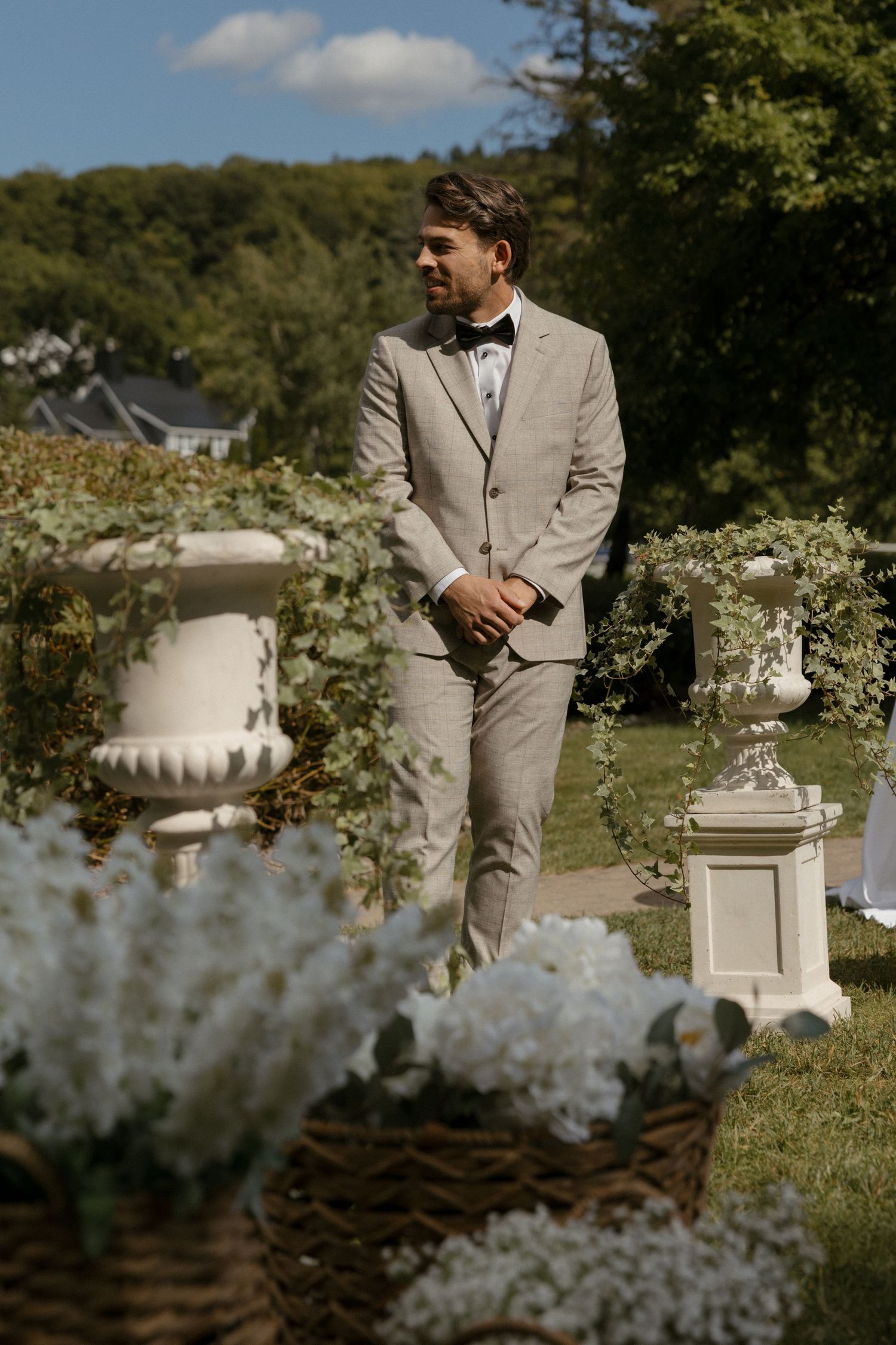 Groom in beige suit with bow tie standing outdoors at garden wedding ceremony surrounded by white flowers and decorative urns.