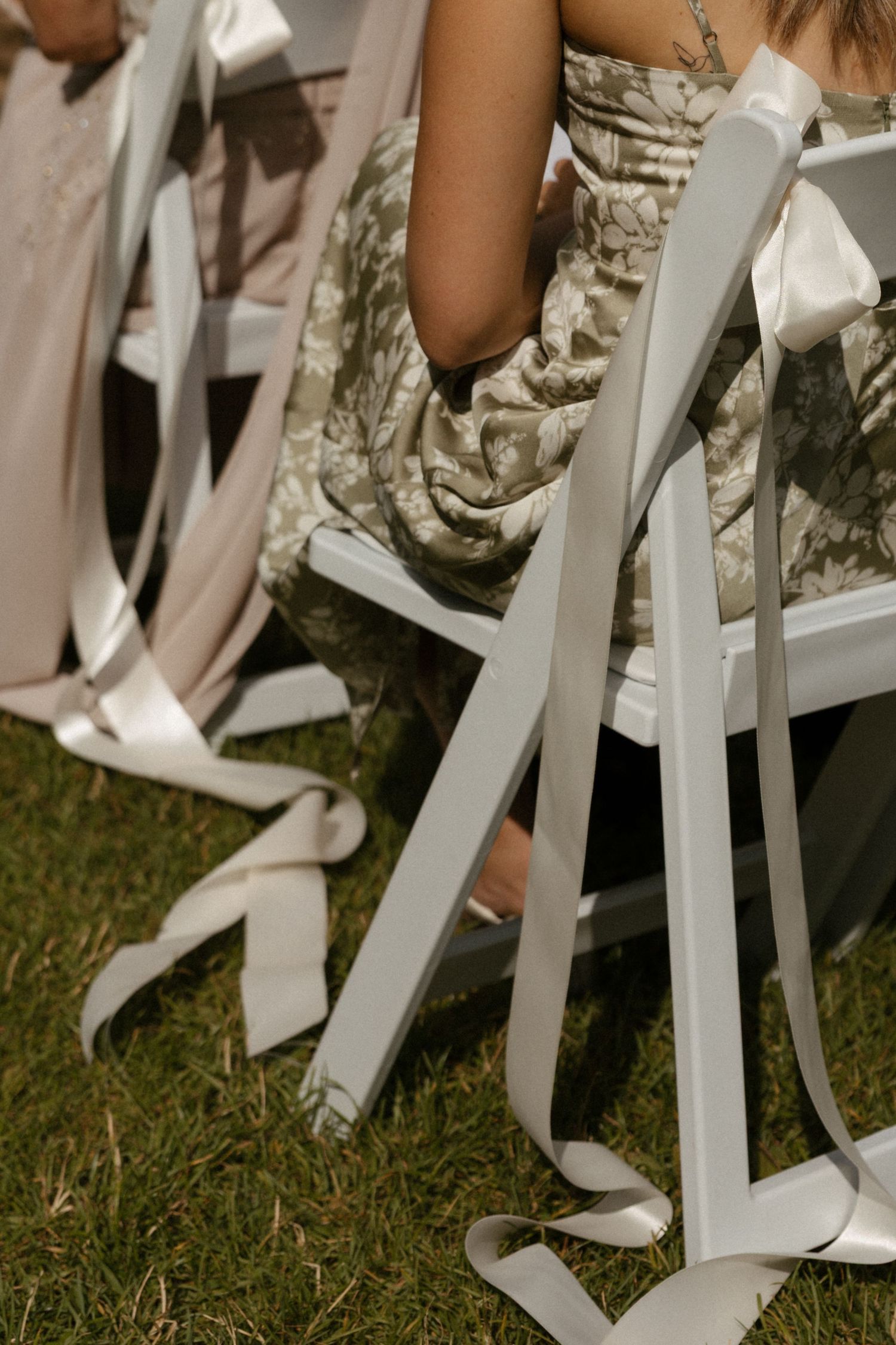 White satin ribbons tied to outdoor wedding chairs with guests in floral dresses seated on green grass.