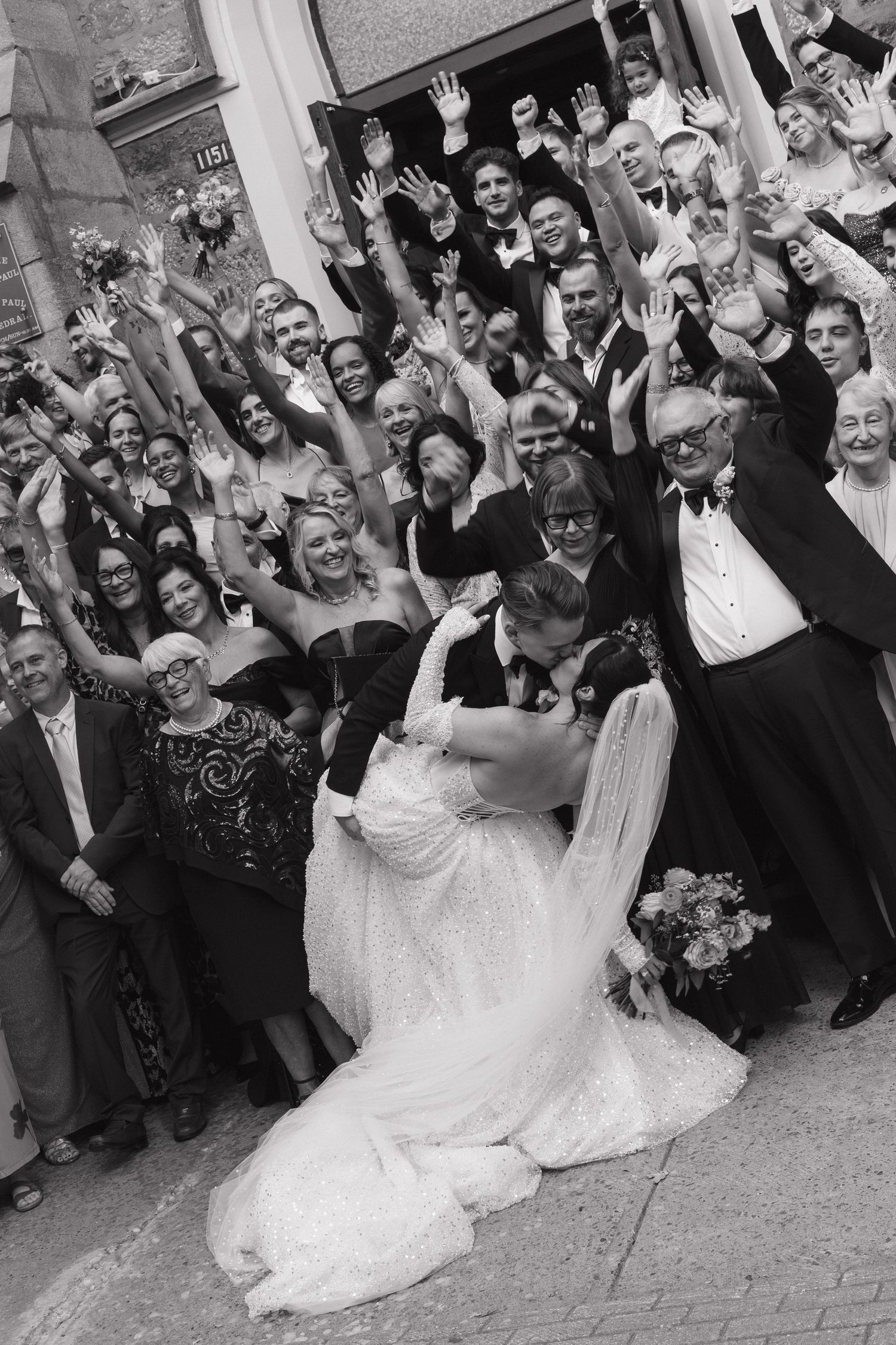 Black and white photo of newlyweds sharing a dip kiss surrounded by cheering wedding guests.