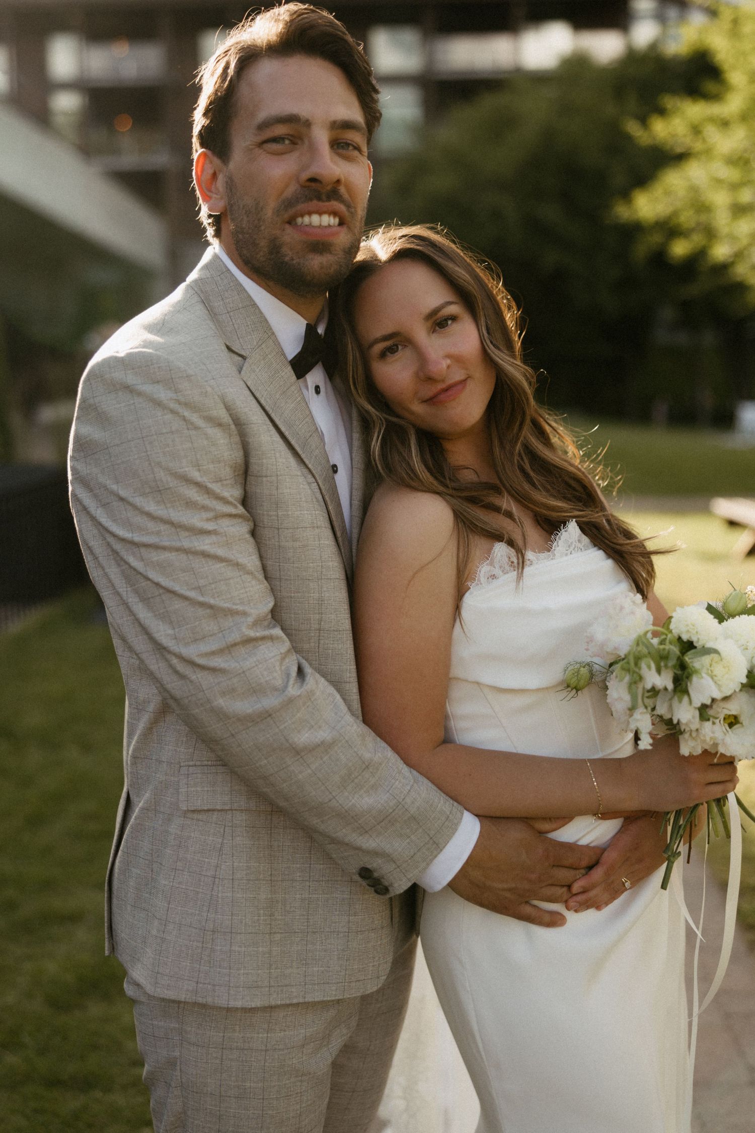 Smiling bride and groom posing outdoors, groom in beige suit embracing bride holding white bouquet.
