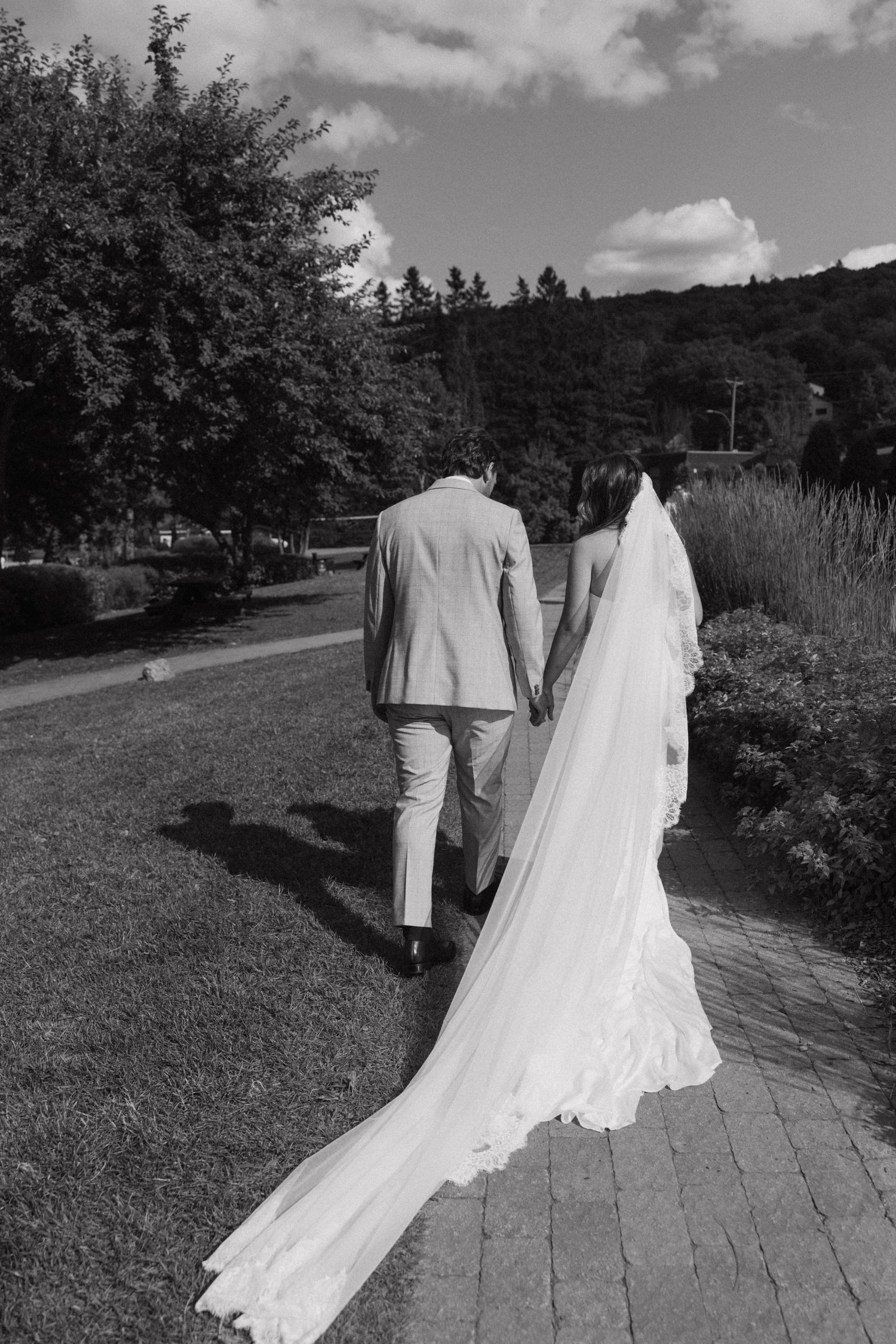 Black and white photo of bride and groom walking hand in hand through a garden path on their wedding day.