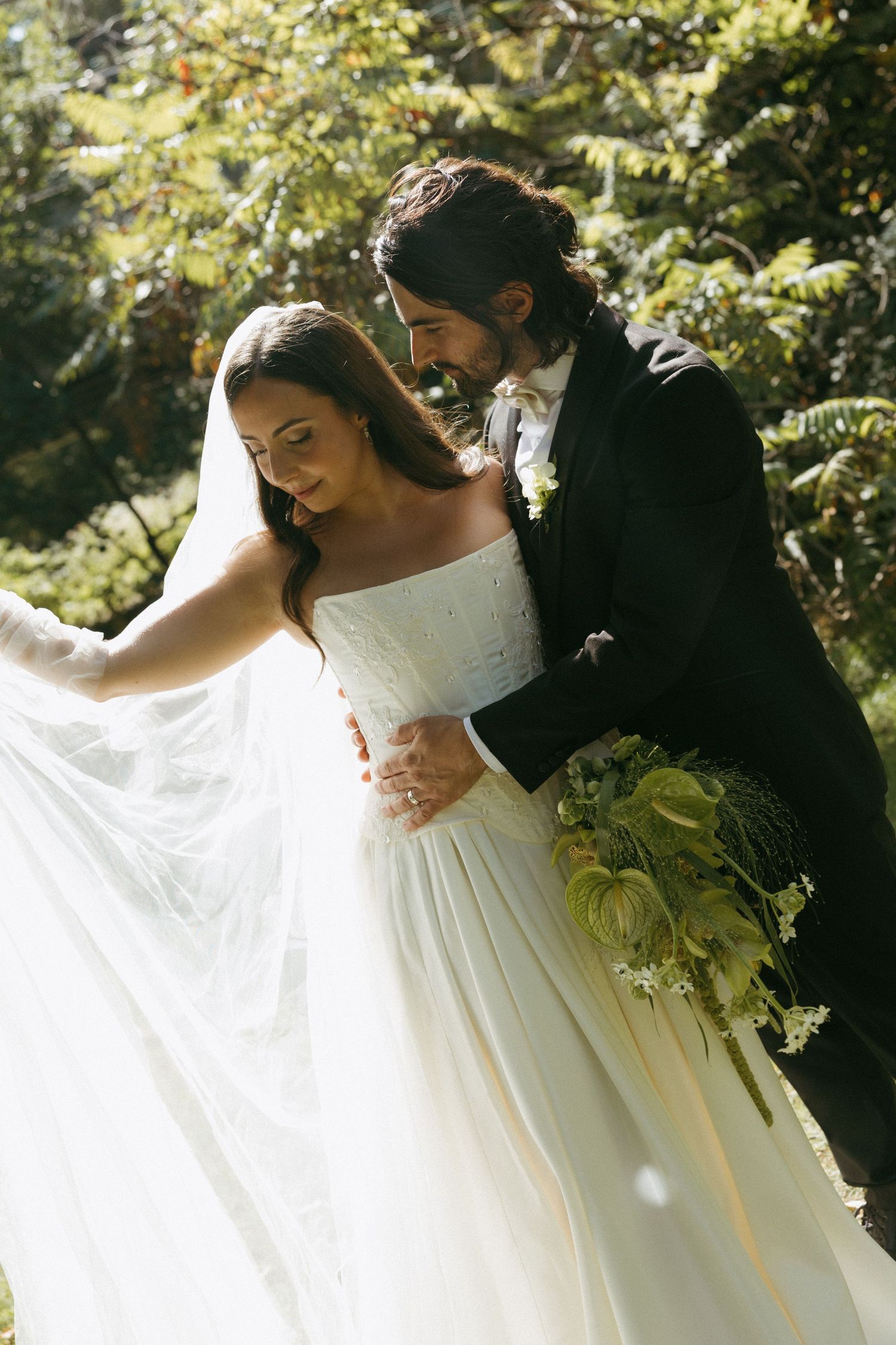 Bride in white gown and groom in black suit share a tender moment outdoors, surrounded by lush green trees.