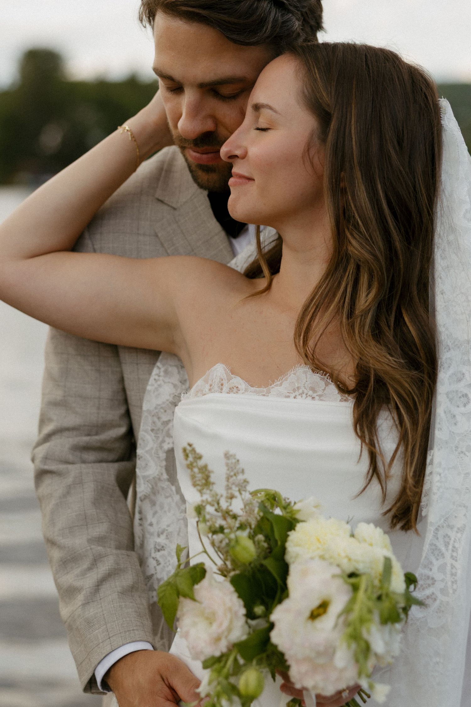A bride holding a white floral bouquet leans into her groom in a beige suit near a lakeside during their wedding.