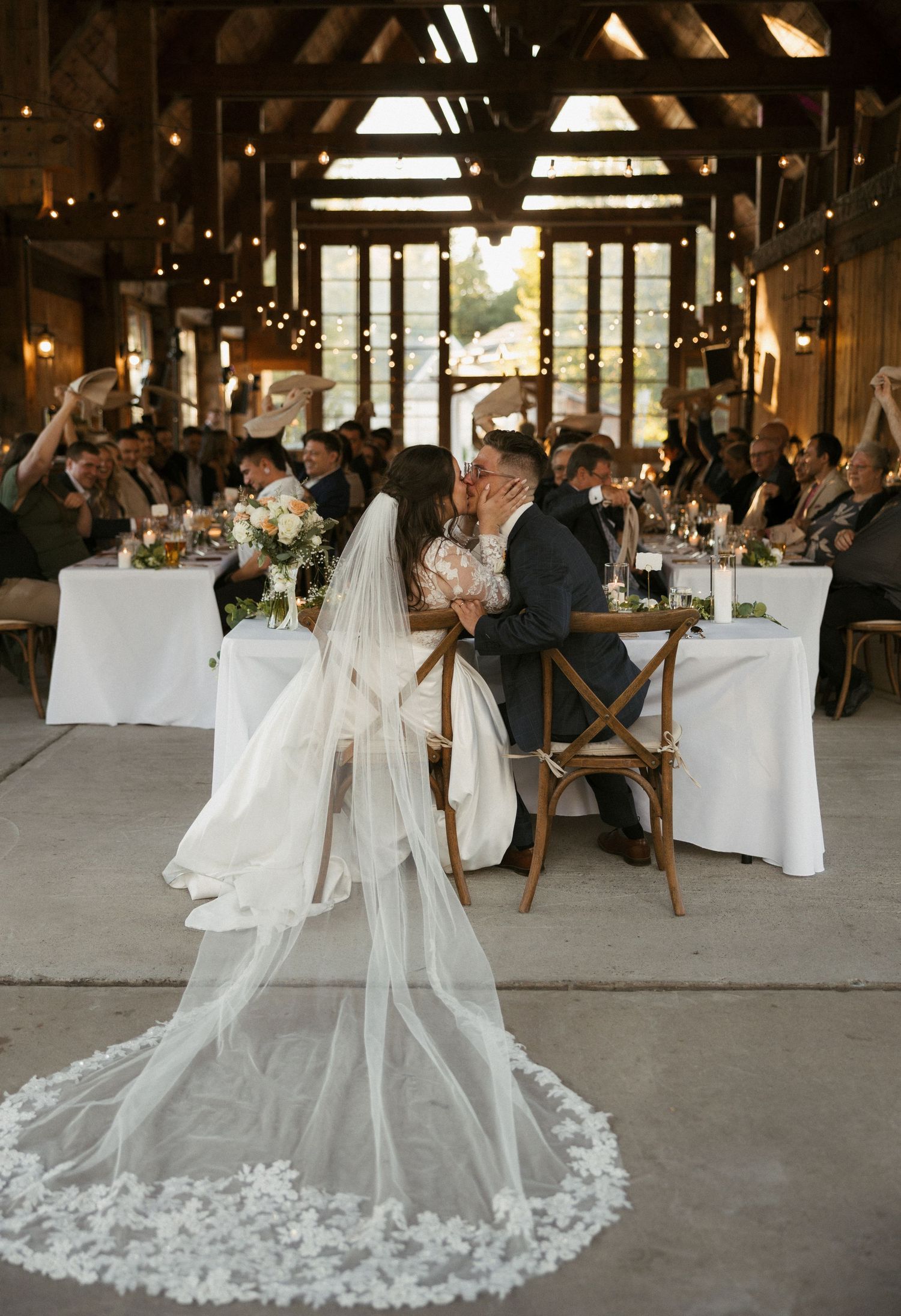 Bride and groom share a kiss at rustic barn wedding reception with lace cathedral veil and cheering guests.