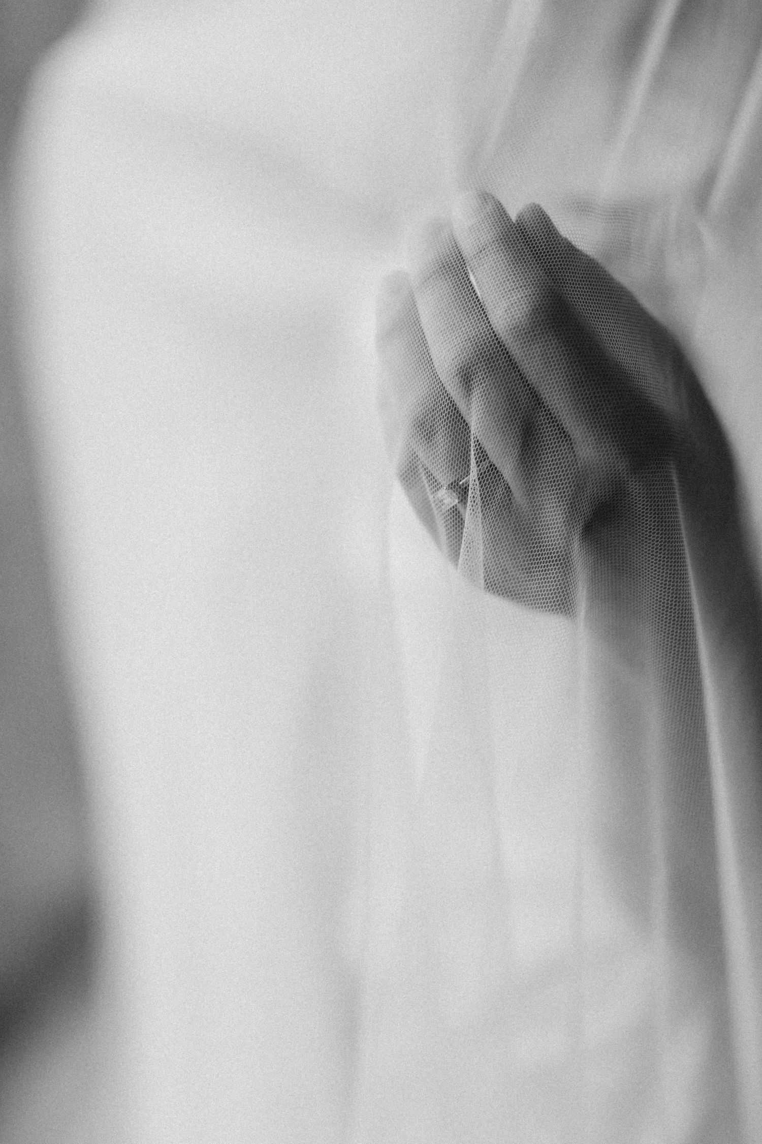 Black and white photo of a hand wearing a delicate ring, gently holding sheer white fabric.