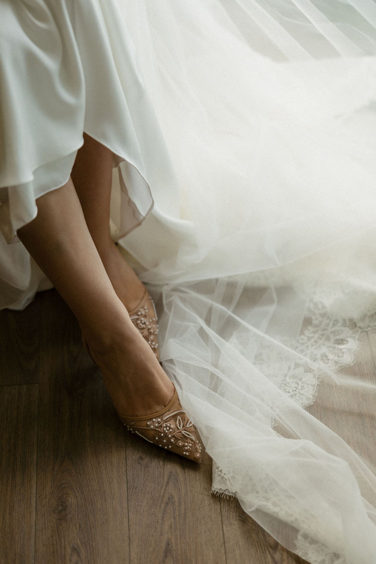 Close-up of a bride wearing embellished gold heels beneath a flowing white wedding dress and lace veil on hardwood floor.
