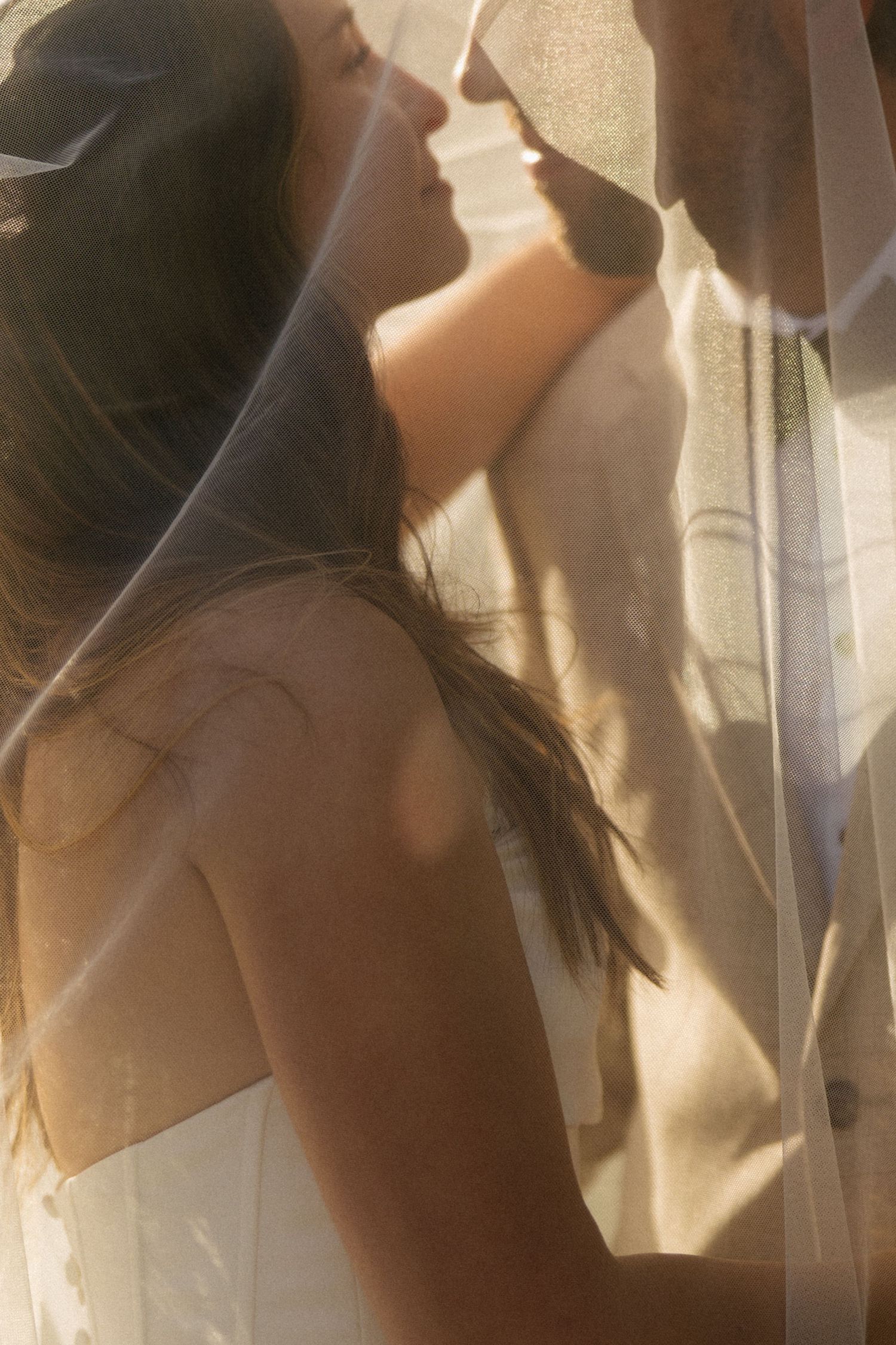 A bride and groom share an intimate moment under a sheer wedding veil in warm golden light.