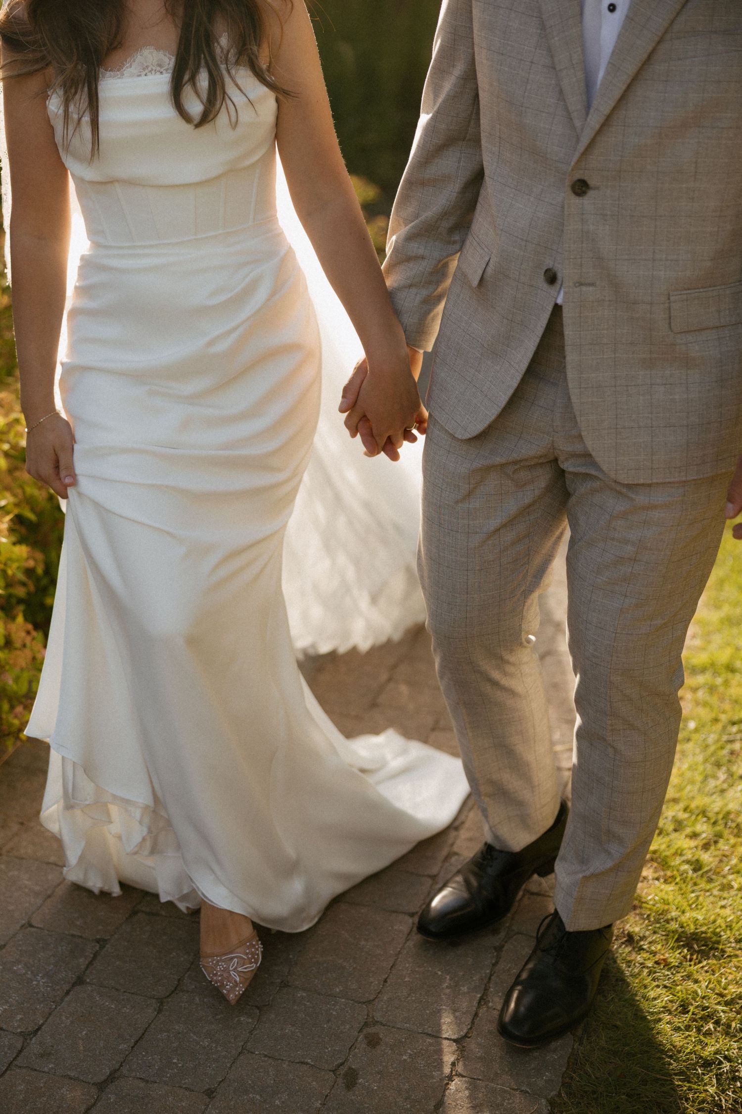 Bride and groom holding hands walking on a brick path, bride in white gown and groom in beige suit.