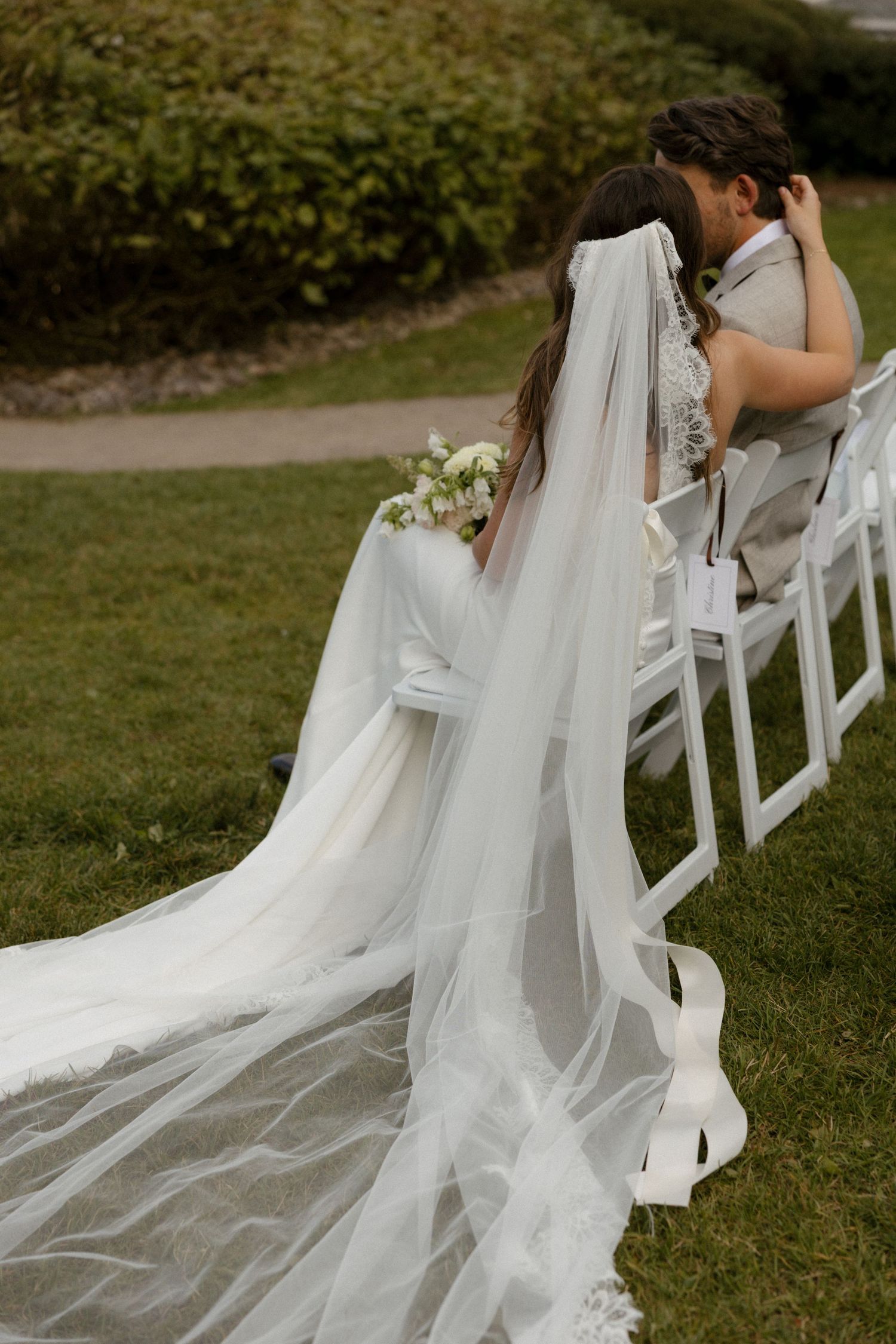 Bride in white gown with long veil holds bouquet seated outdoors at garden wedding ceremony.