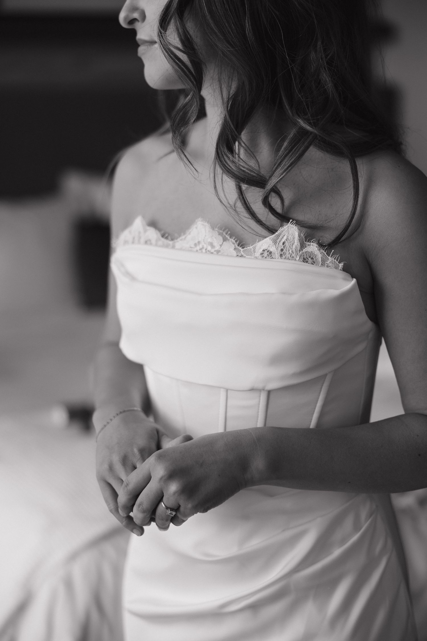 Black and white photo of a bride in a strapless lace wedding gown, hands clasped, wearing a ring.
