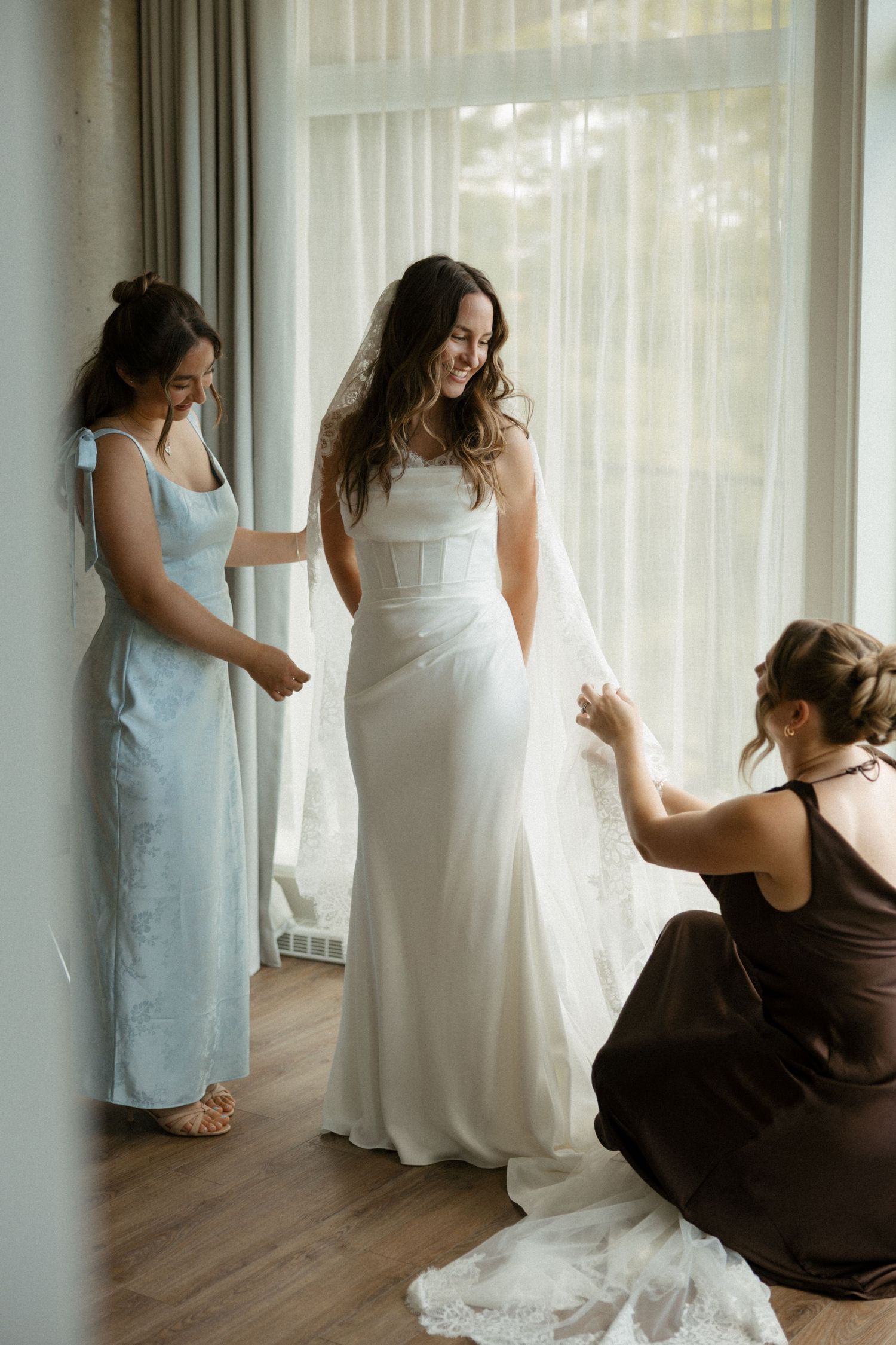 Bridesmaids in blue and brown dresses help smiling bride into elegant white wedding gown near bright window.