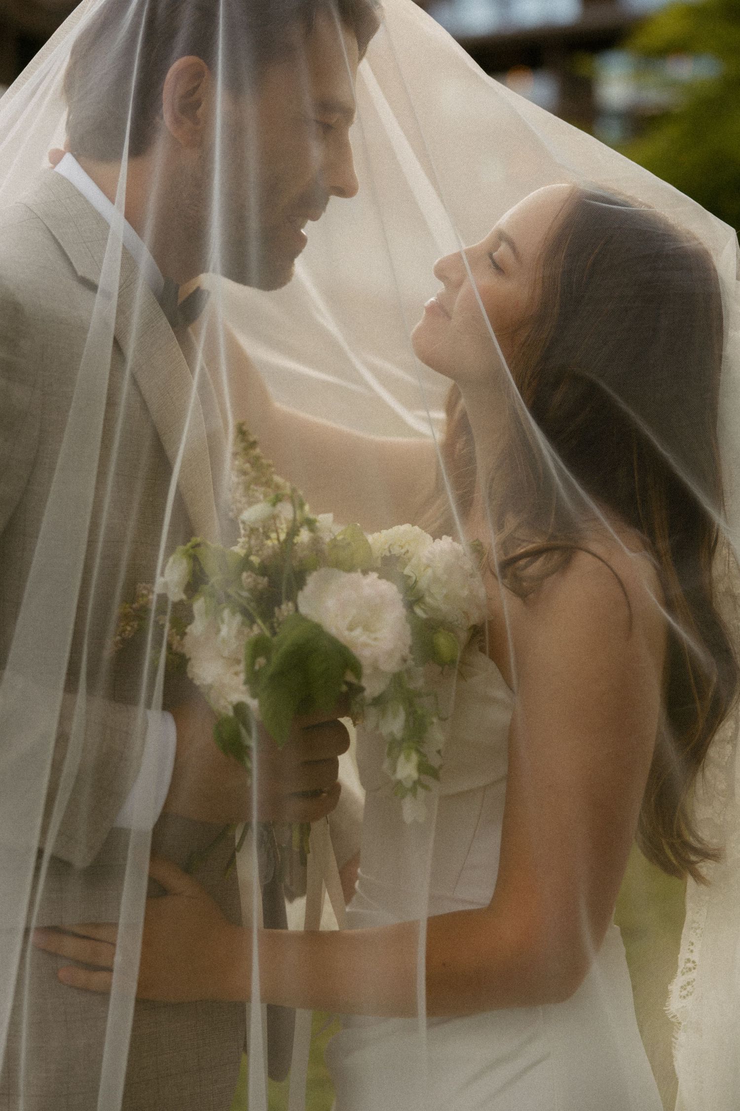 Bride and groom share an intimate moment under a flowing white veil, bride holding a white floral bouquet.