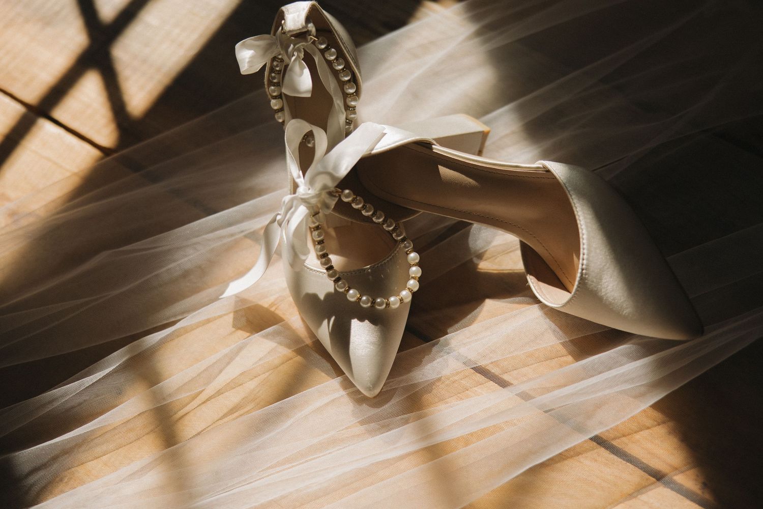 Elegant bridal heels with pearl bracelet and satin bow on wooden floor with soft sunlight and flowing veil.