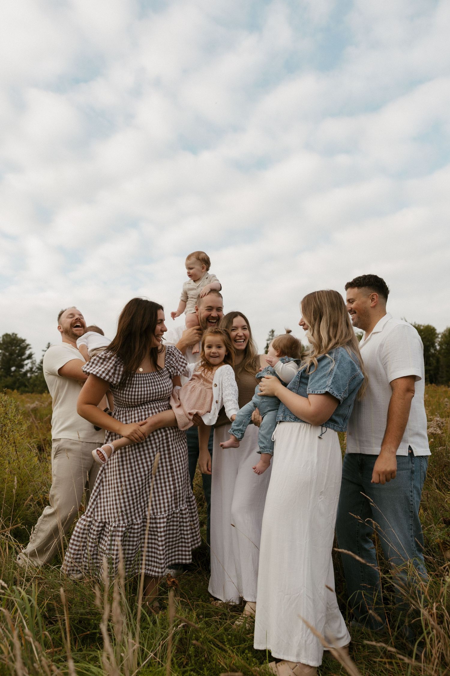 Large family group smiling and laughing together in a sunny field, holding babies and toddlers, dressed in neutral tones.