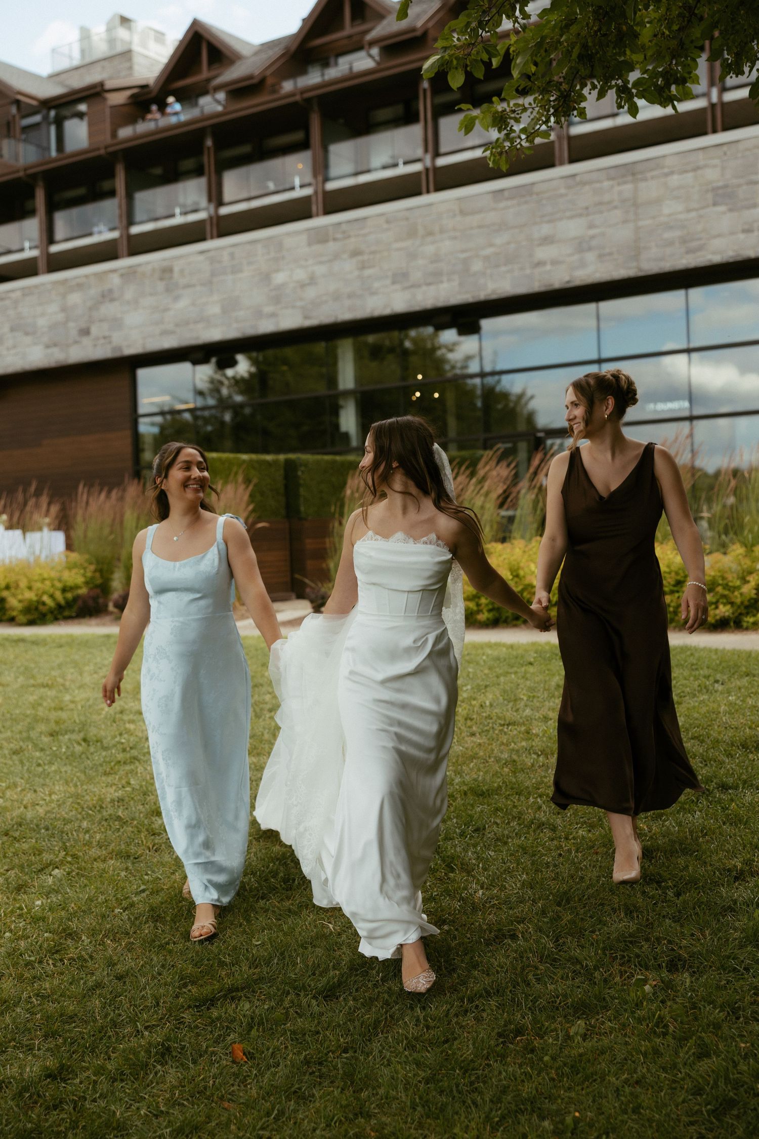 Bride in white gown walks with two bridesmaids in light blue and dark dresses on green lawn outside modern venue.