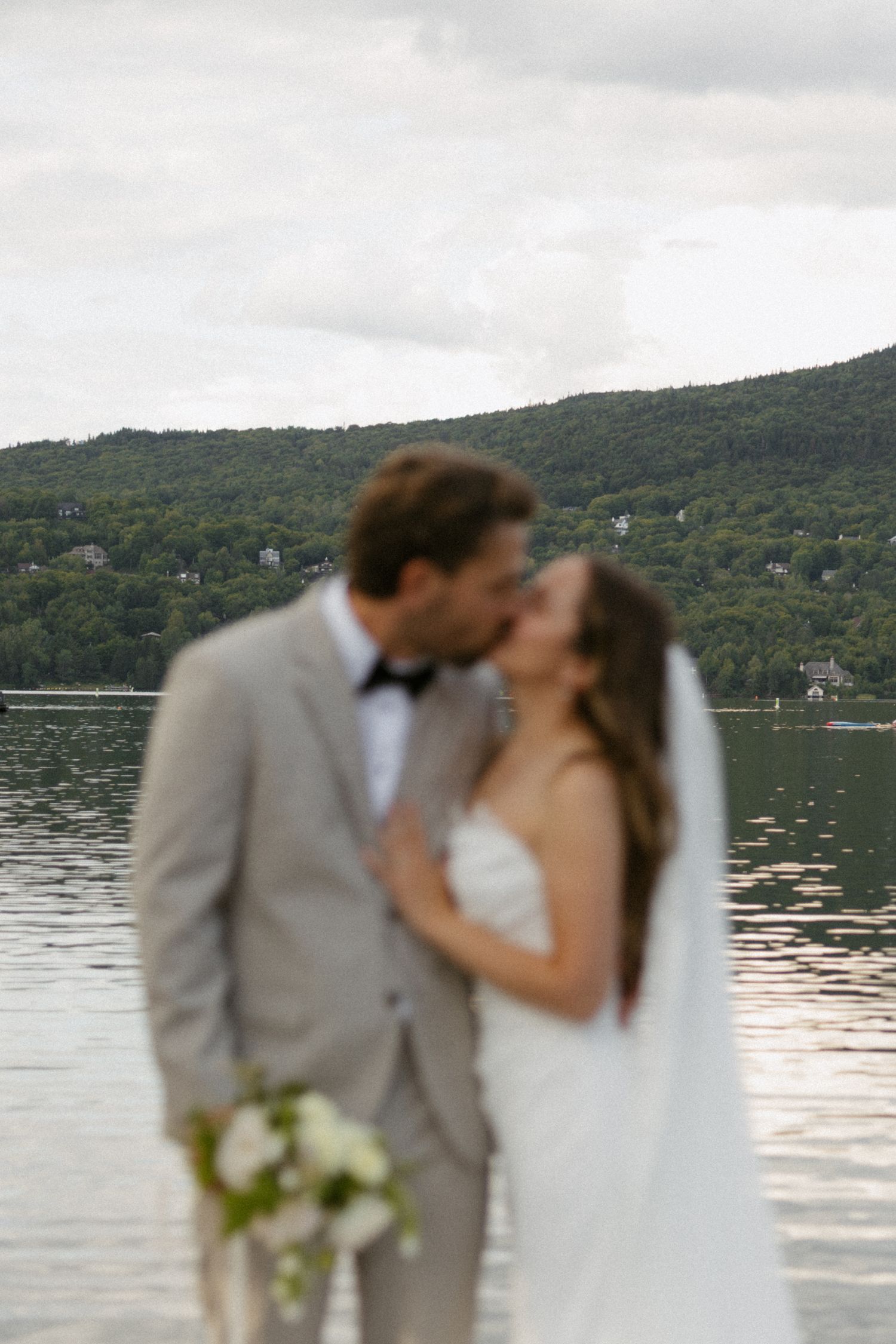 A bride and groom share a romantic kiss by a serene lake with lush green hills in the background on their wedding day.