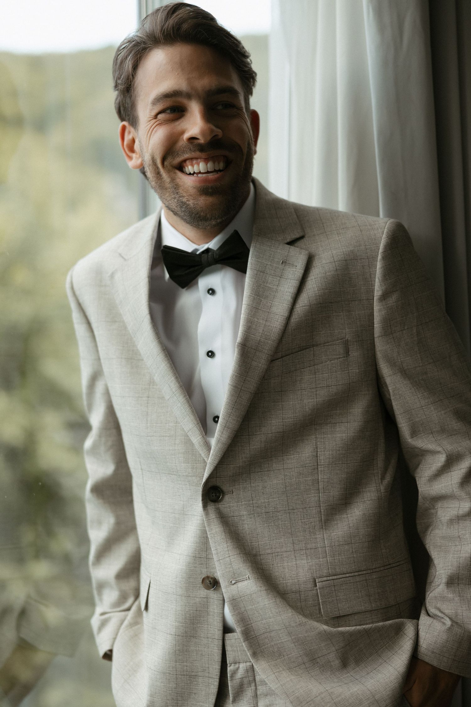 Smiling groom in light gray tuxedo jacket with black bow tie standing by a window on his wedding day.