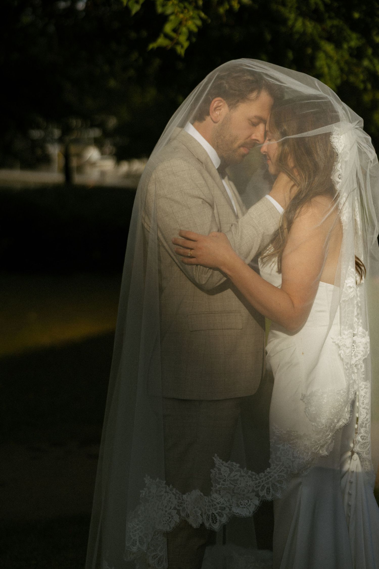 Bride and groom share romantic moment under lace wedding veil in golden outdoor light.
