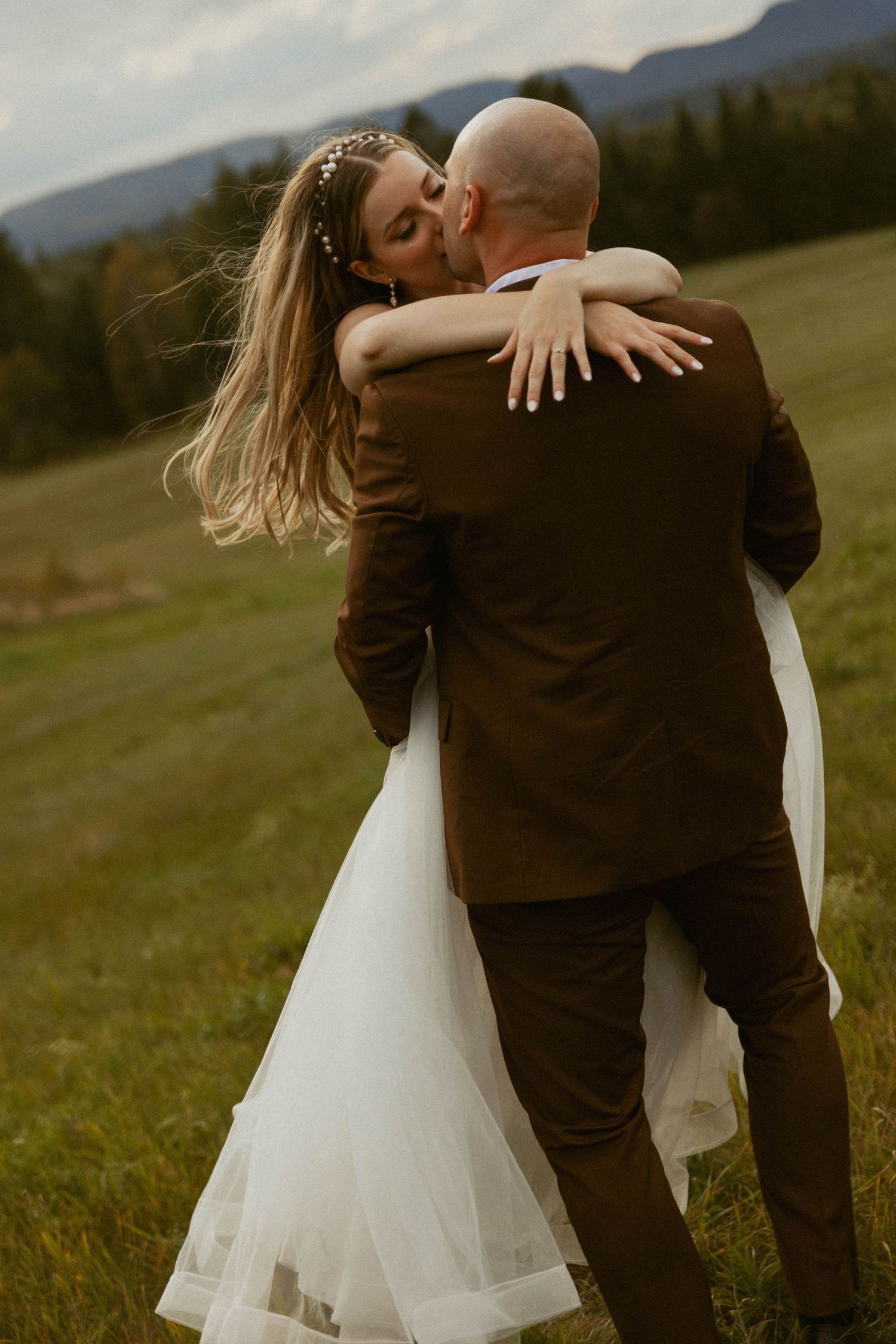 A bride in a white dress kisses her bald groom in a brown suit outdoors in a scenic mountain meadow.