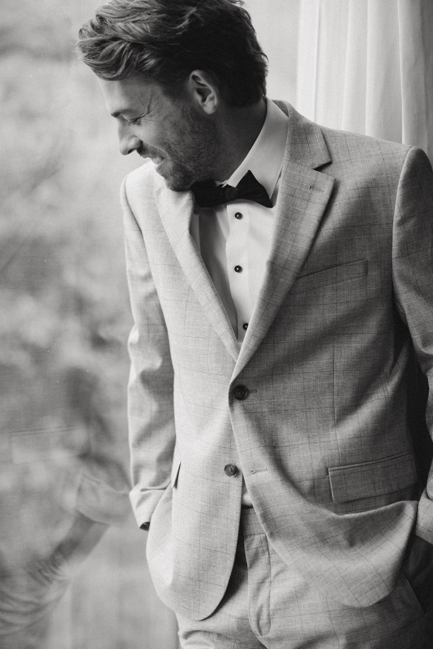 Black and white photo of a smiling groom in a light suit and bow tie standing by a window.