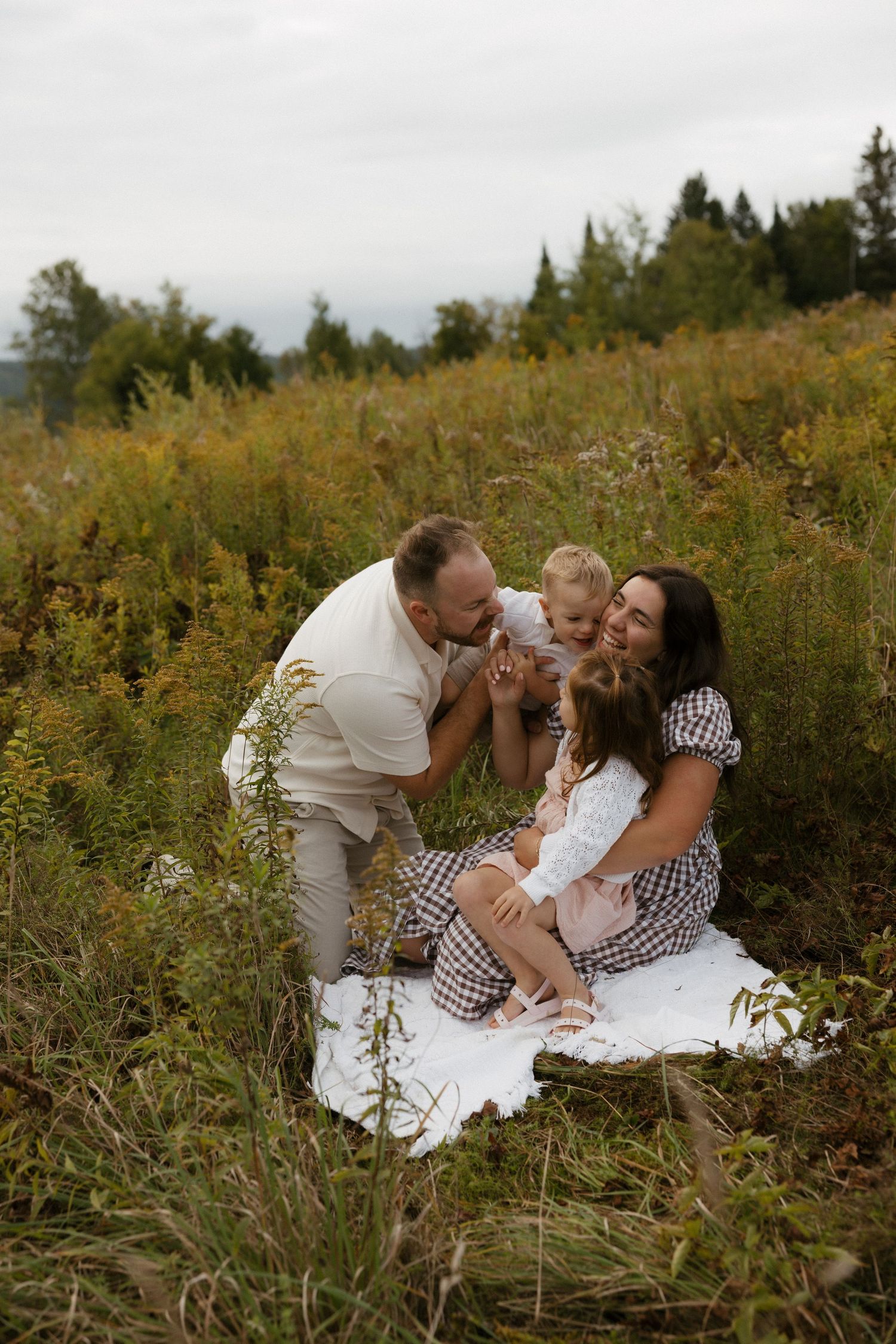 Family of four enjoying a picnic in a wildflower meadow, parents kissing baby while daughter sits on blanket.