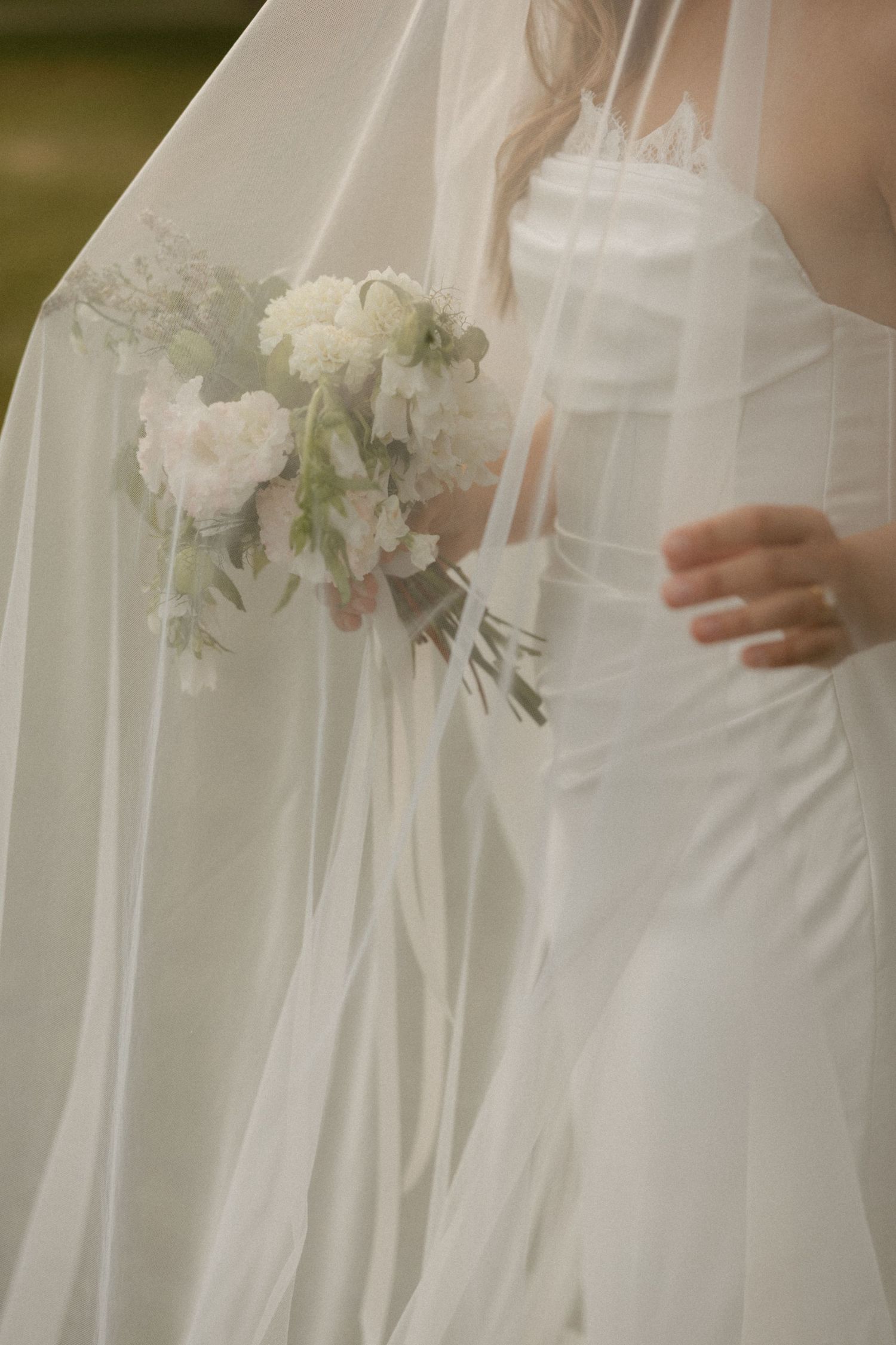 Bride in white strapless gown holding delicate white floral bouquet, draped with sheer veil outdoors.
