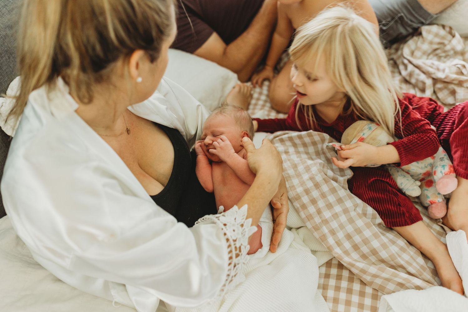 Mother holds newborn baby while young girl in red pajamas gently touches the infant on a white bed.